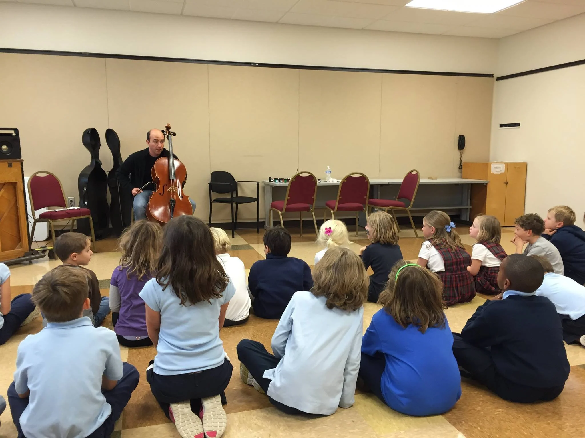 A man with a cello performing for a group of elementary school children sitting on the floor in a classroom or auditorium.