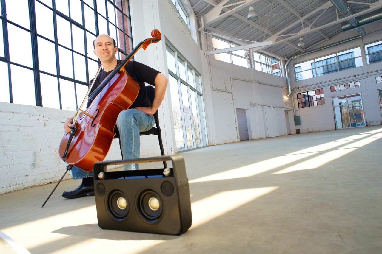 A man sitting on a chair playing a cello inside a spacious industrial-style room with large windows and exposed ceiling beams. A portable speaker is placed on the floor nearby.