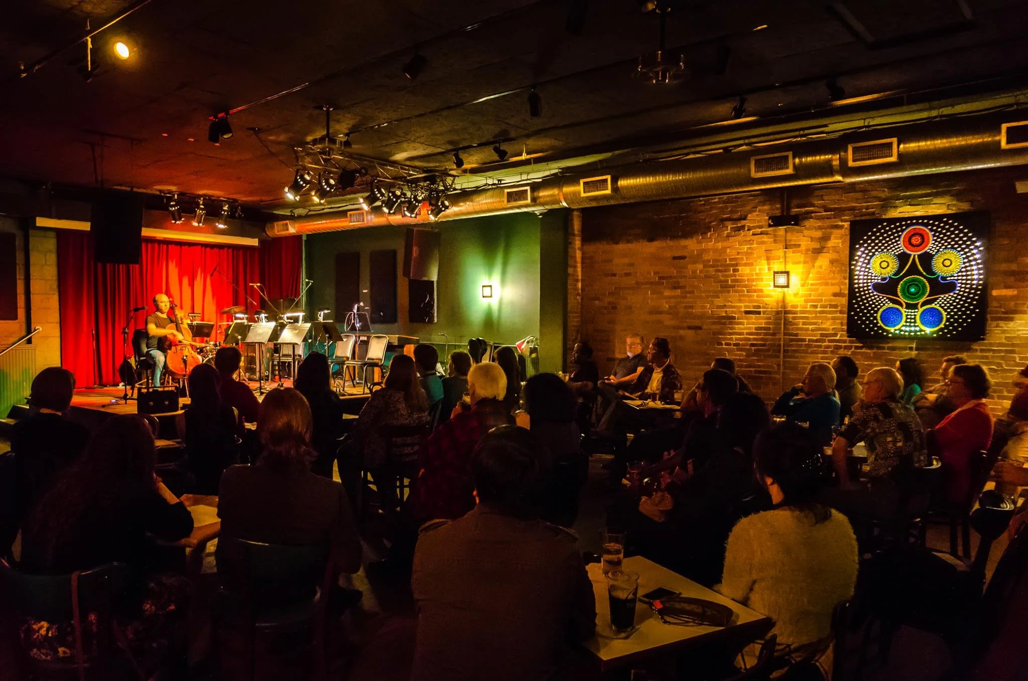 An indoor jazz concert with a musician playing the cello on stage, audience seated in front, red curtains and wall art, warm lighting, brick wall.