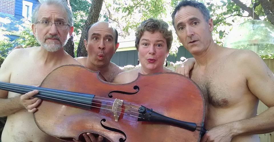Four shirtless men standing outdoors behind a tree, holding a large stringed instrument, with trees and a fence in the background, looking at the camera with surprised expressions.