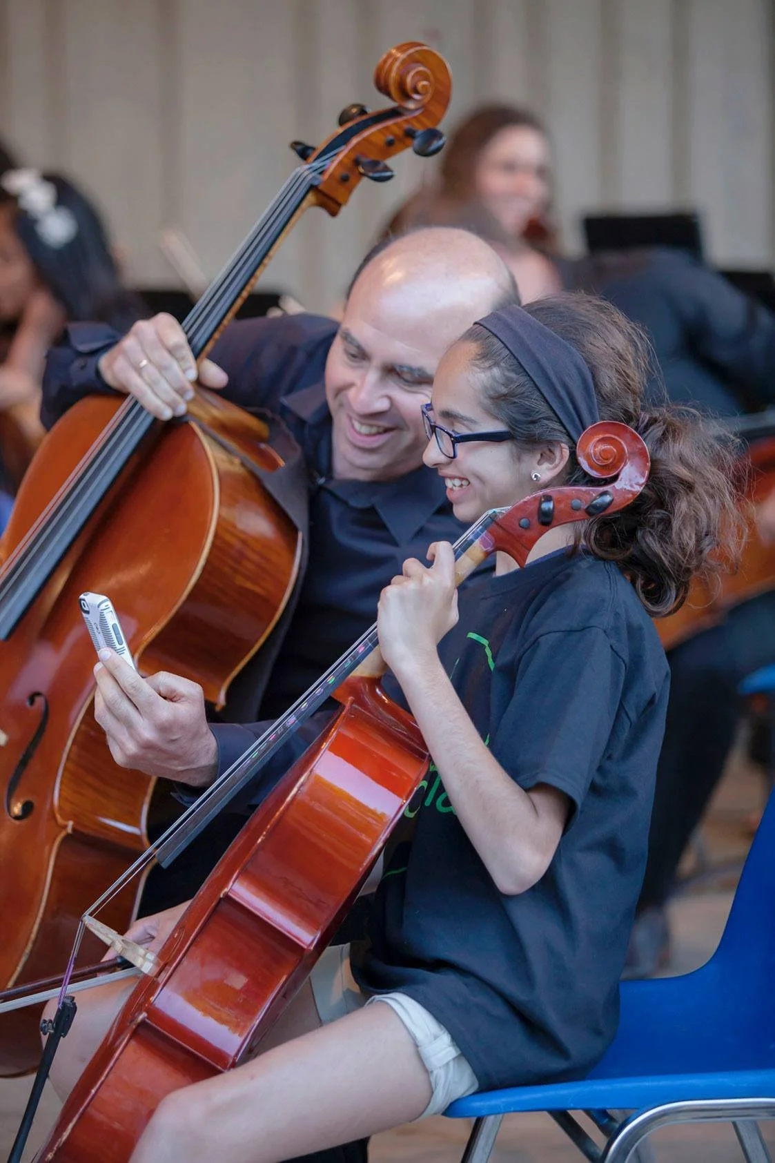 A man and a girl are sitting together, both playing cellos and smiling while looking at a device.
