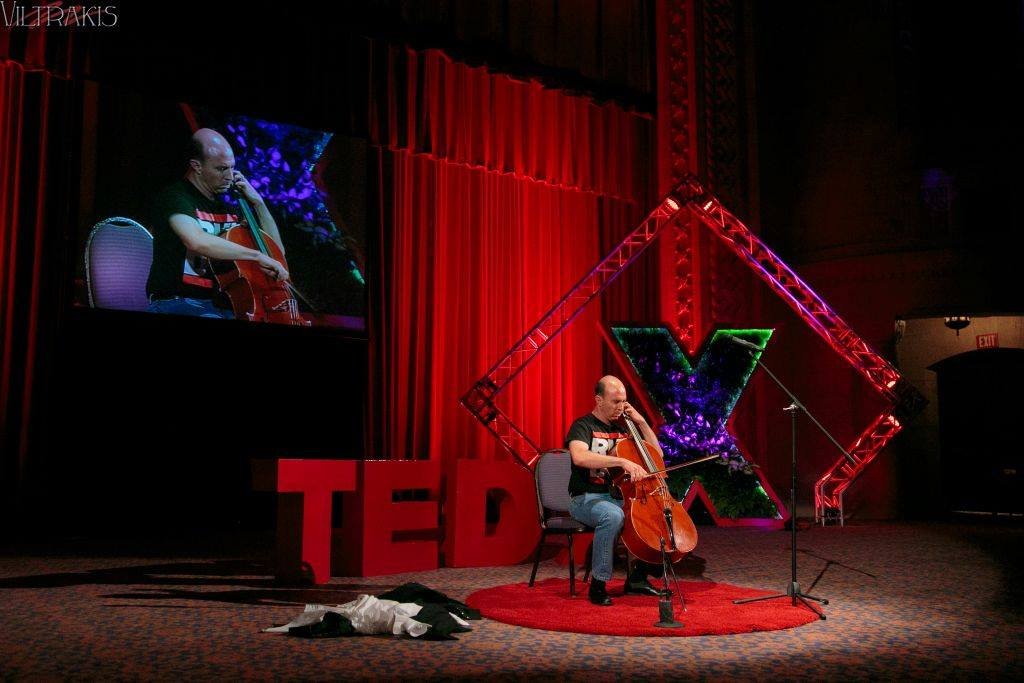 A man playing a cello on a stage with red curtains and TEDx branding, including large red 3D letters spelling 'TED', a vertical lights structure, and a person’s image projected on a screen next to him.