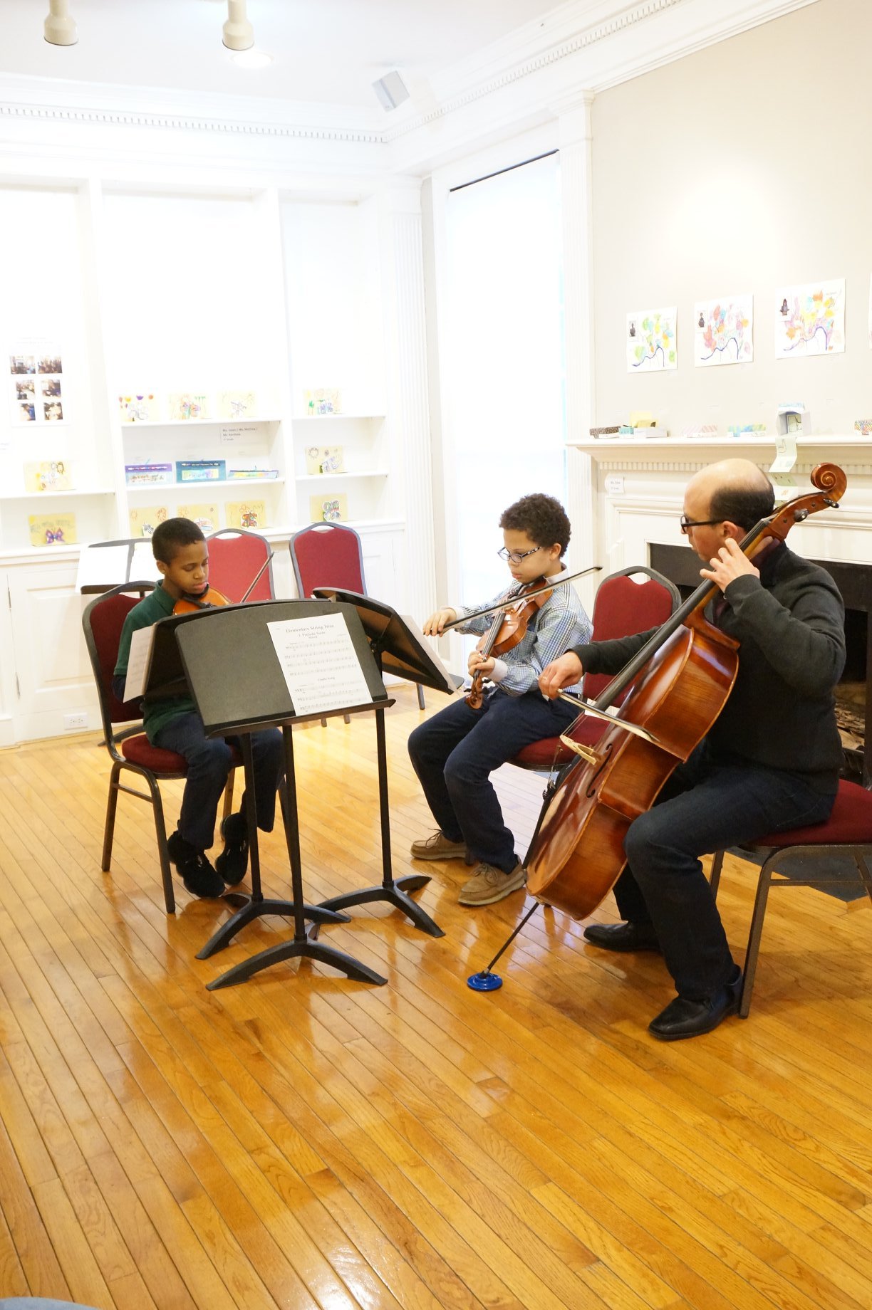 Three musicians, two children and an adult, playing string instruments in a bright room with wooden floors and artwork on the walls.