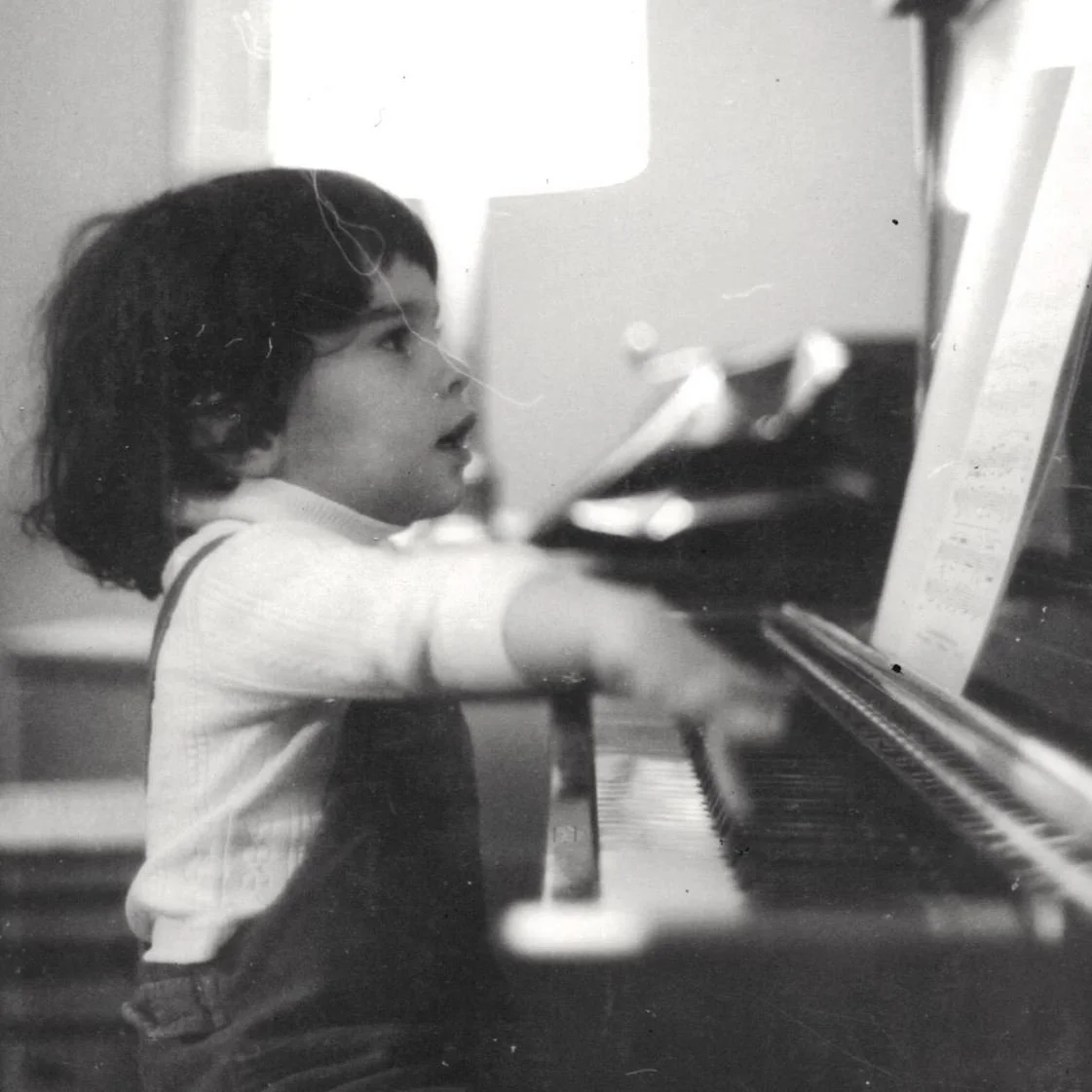 A young child with dark hair playing the piano, appearing to be focused on the keys, in a room with a lamp and some furniture.