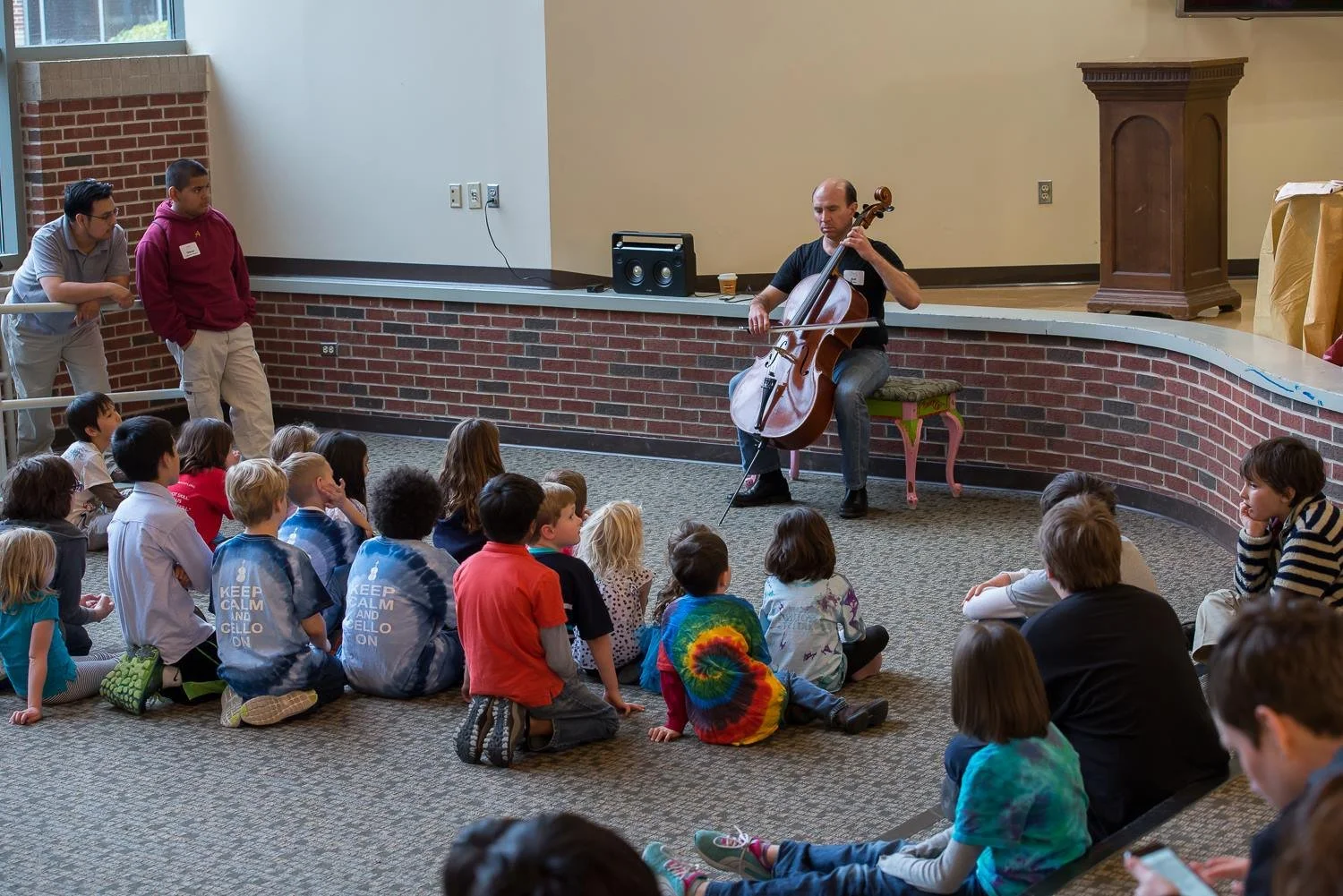 A man playing a cello in front of a group of children sitting on the carpeted floor, with two adults standing nearby watching. The setting appears to be an indoor venue with a curved brick wall and podium in the background.