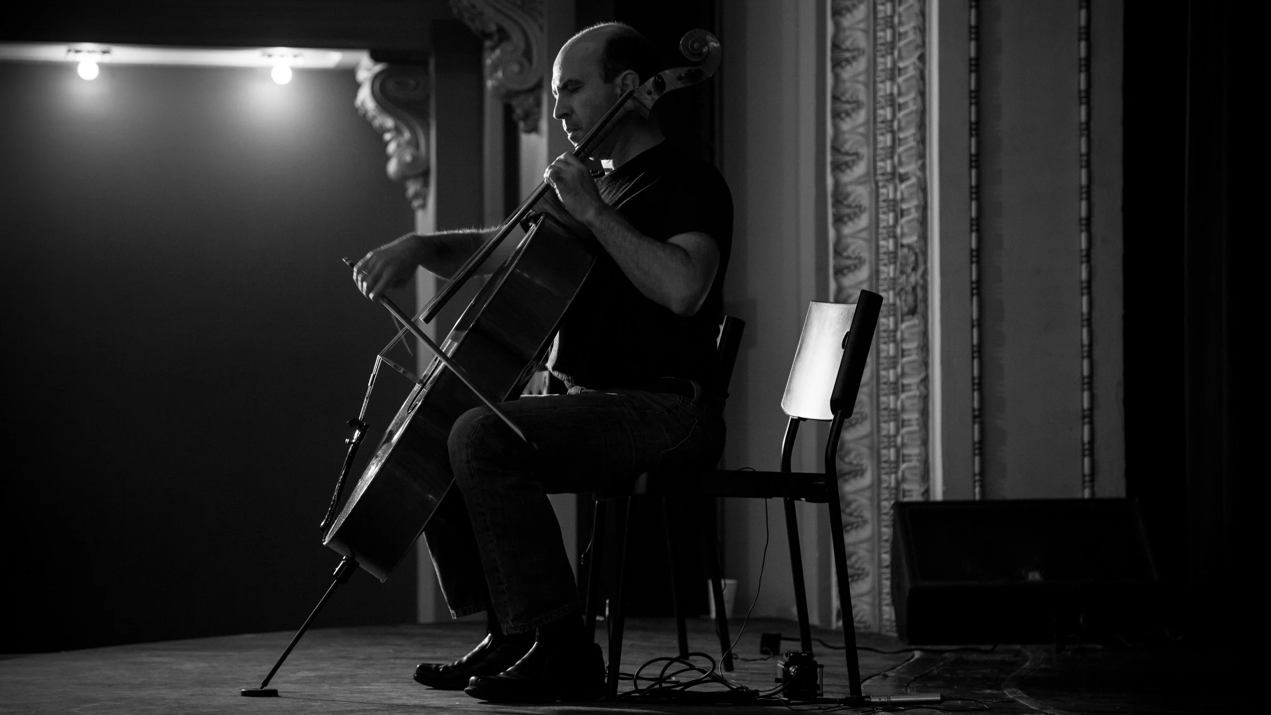 A man playing the cello on stage in a black-and-white photo, sitting on a chair with a music stand nearby, in an ornate room with decorative molding.