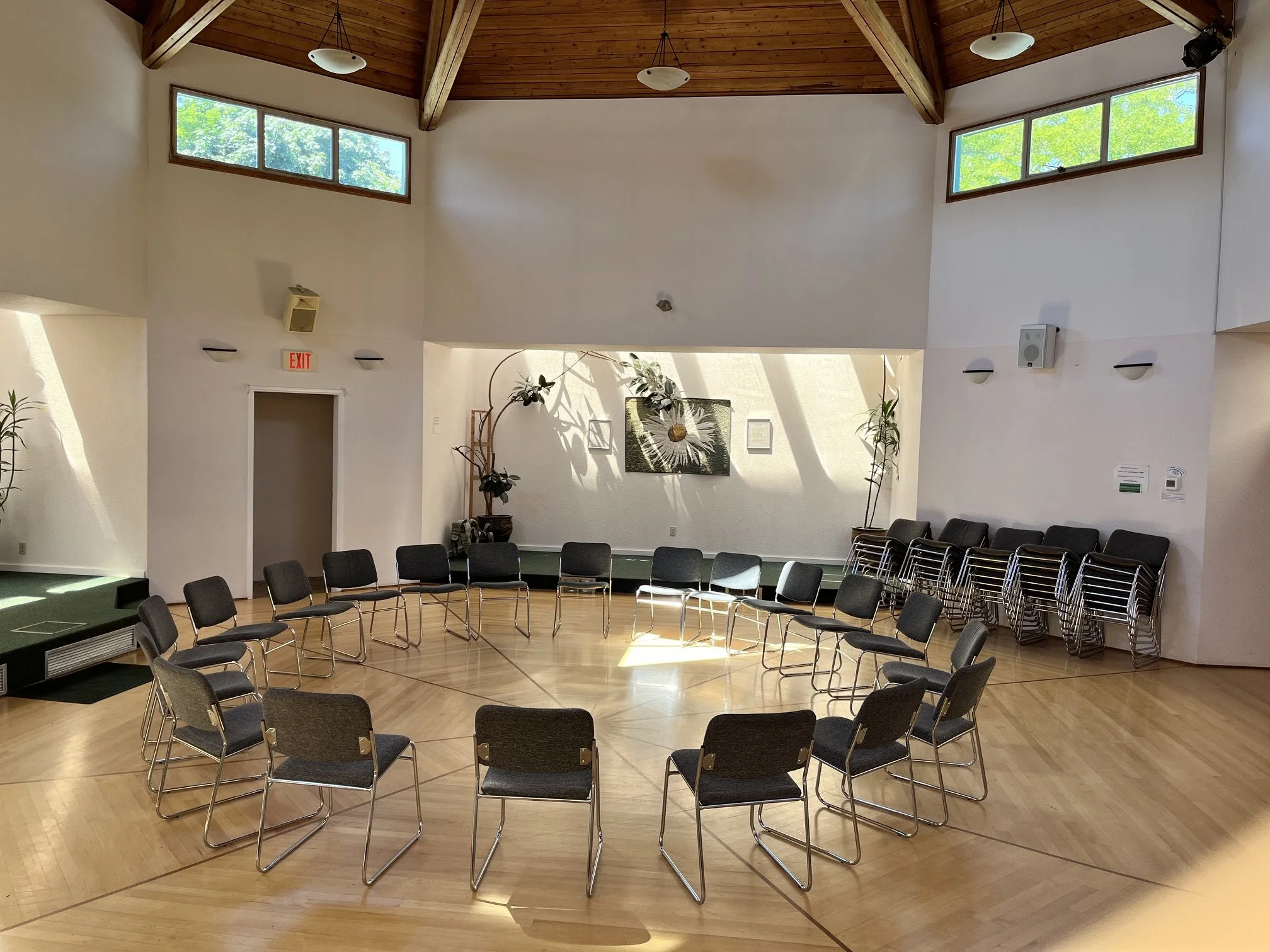 Interior of a room with a high wooden ceiling, multiple windows, and a semi-circular arrangement of black chairs. There are potted plants, framed artwork, and a blank wall.
