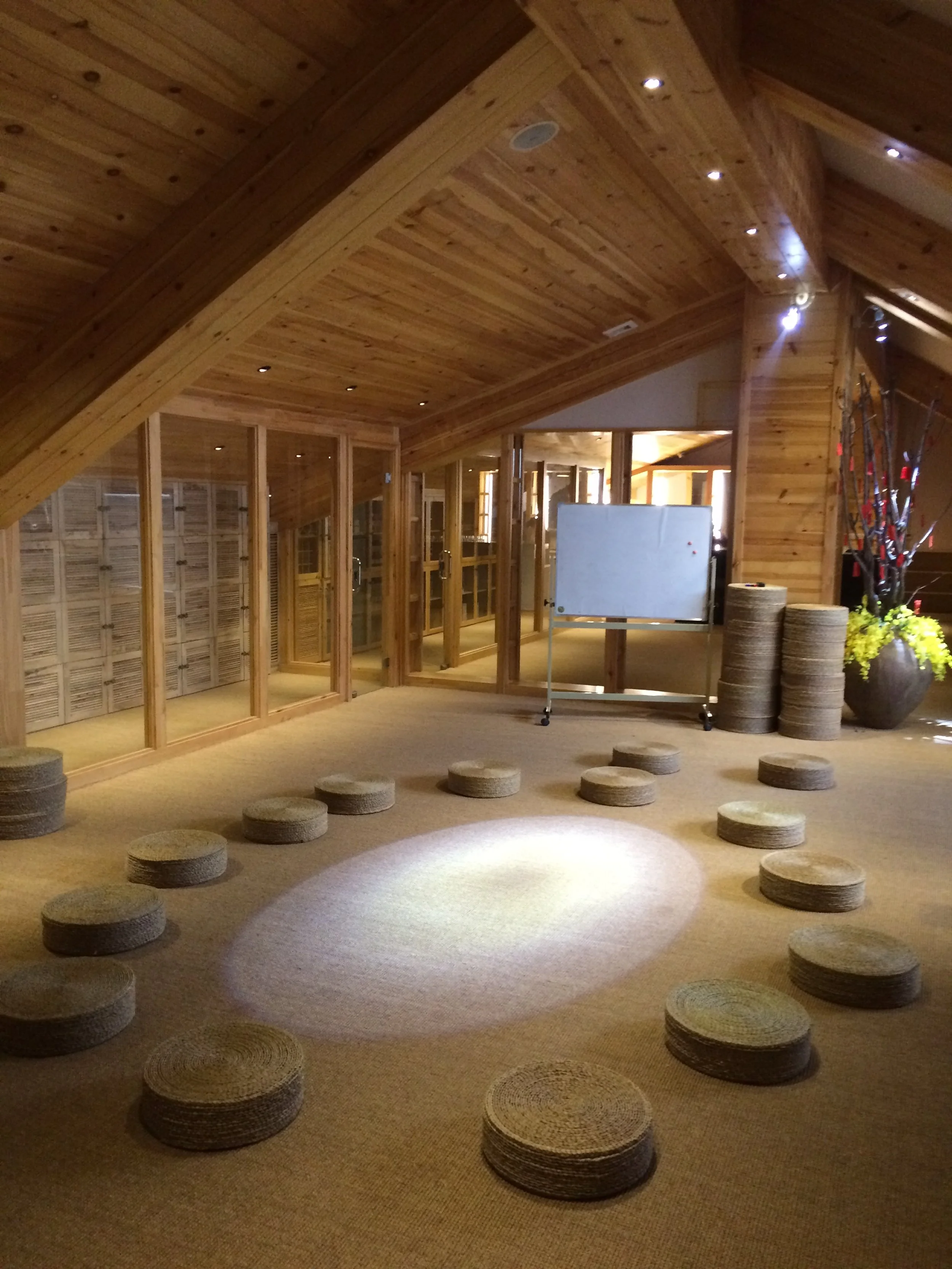 Interior of a wooden room with a sloped ceiling, glass door walls, and round woven mats on the carpeted floor, some arranged in a pathway and others in piles. There is a whiteboard and decorative branches with red ornaments and yellow flowers.