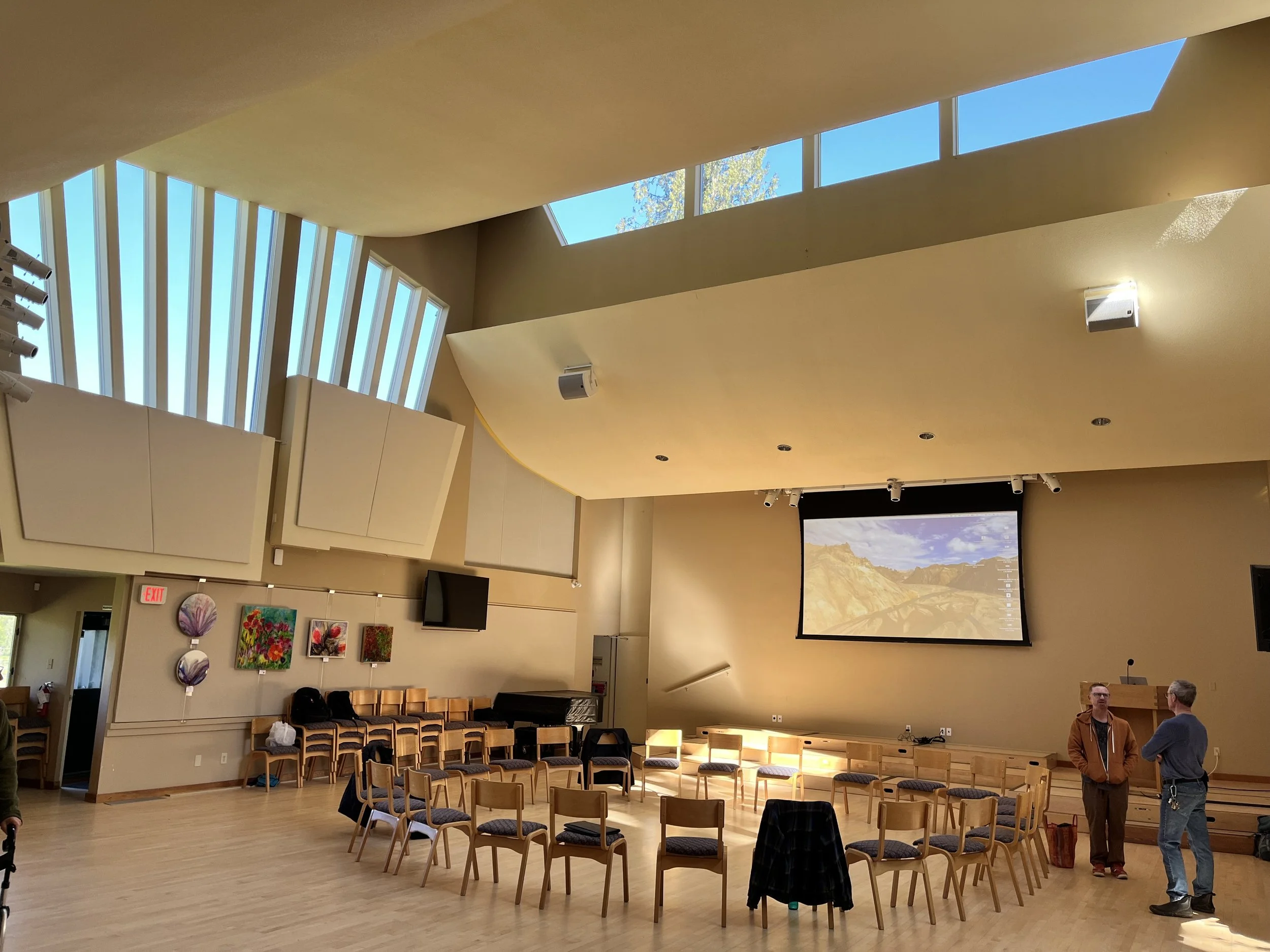Interior of a large room with rows of wooden chairs, a large overhead screen displaying desert landscape, and a few people standing and talking.