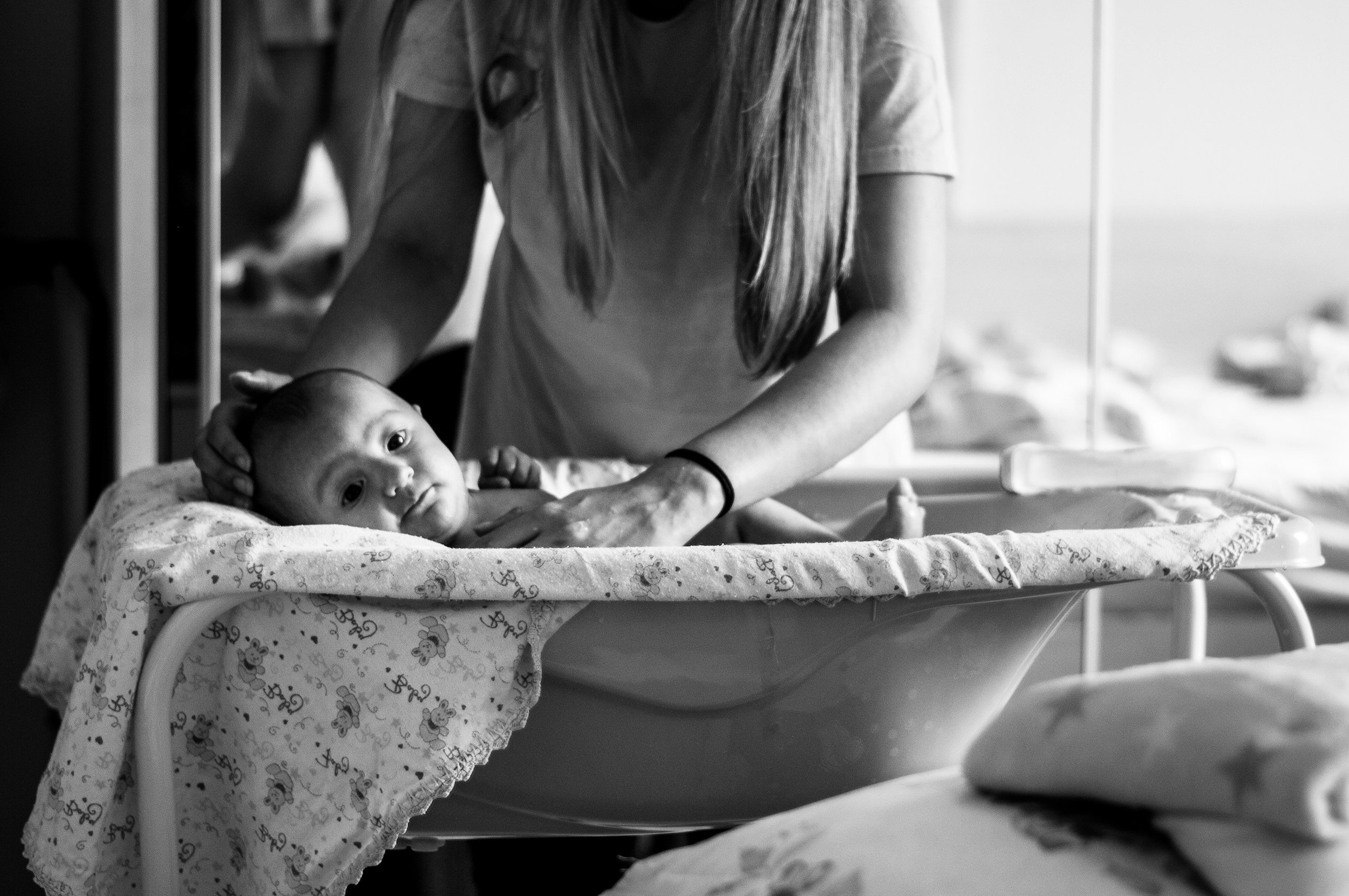 A woman in a hospital or medical setting gently holds a newborn baby in an infant hospital bassinet, with a view of a window showing a blurry outdoor scene.