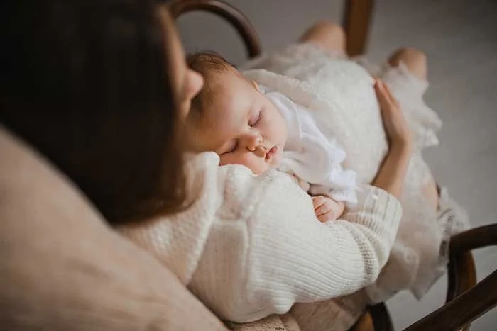 A woman holding a sleeping baby in her arms, sitting on a wooden chair.