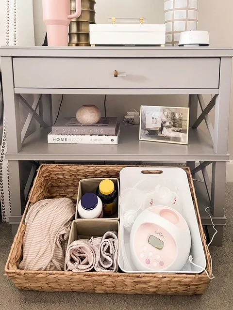 A wicker basket on the floor containing baby items such as a striped onesie, baby socks, bottles of vitamins or supplements, and a baby scale. The basket is placed in front of a gray shelf with decor pieces on top, including vases, a framed photo, and books.