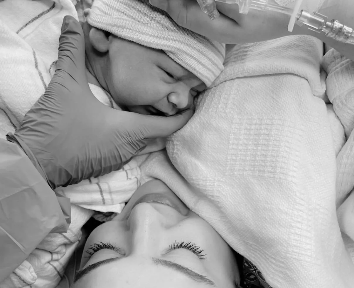 A newborn baby wearing a hat and wrapped in a blanket, sleeping on a hospital bed, with a person touching its face gently.