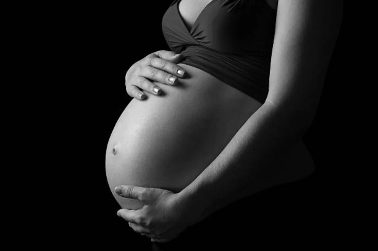 Black and white photo of a pregnant woman with her hands on her belly, wearing a dark top, against a dark background.