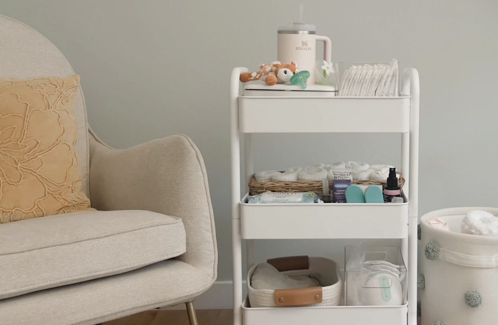 A white multi-tiered changing table filled with baby products, next to a beige couch with a decorative pillow, and a laundry basket with towels on a light-colored wall.