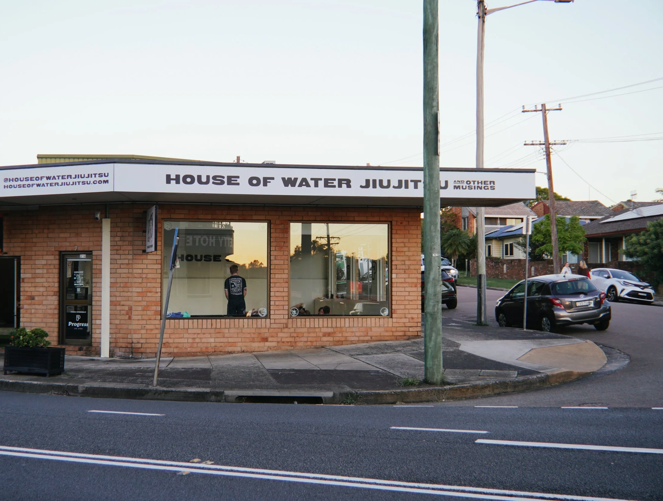 A brick building with a sign reading 'House of Water Jiu Jitsu' on the roof. Large windows show people inside, with one person visible standing near the window. Several cars are parked along the street and residential houses are in the background at sunset.