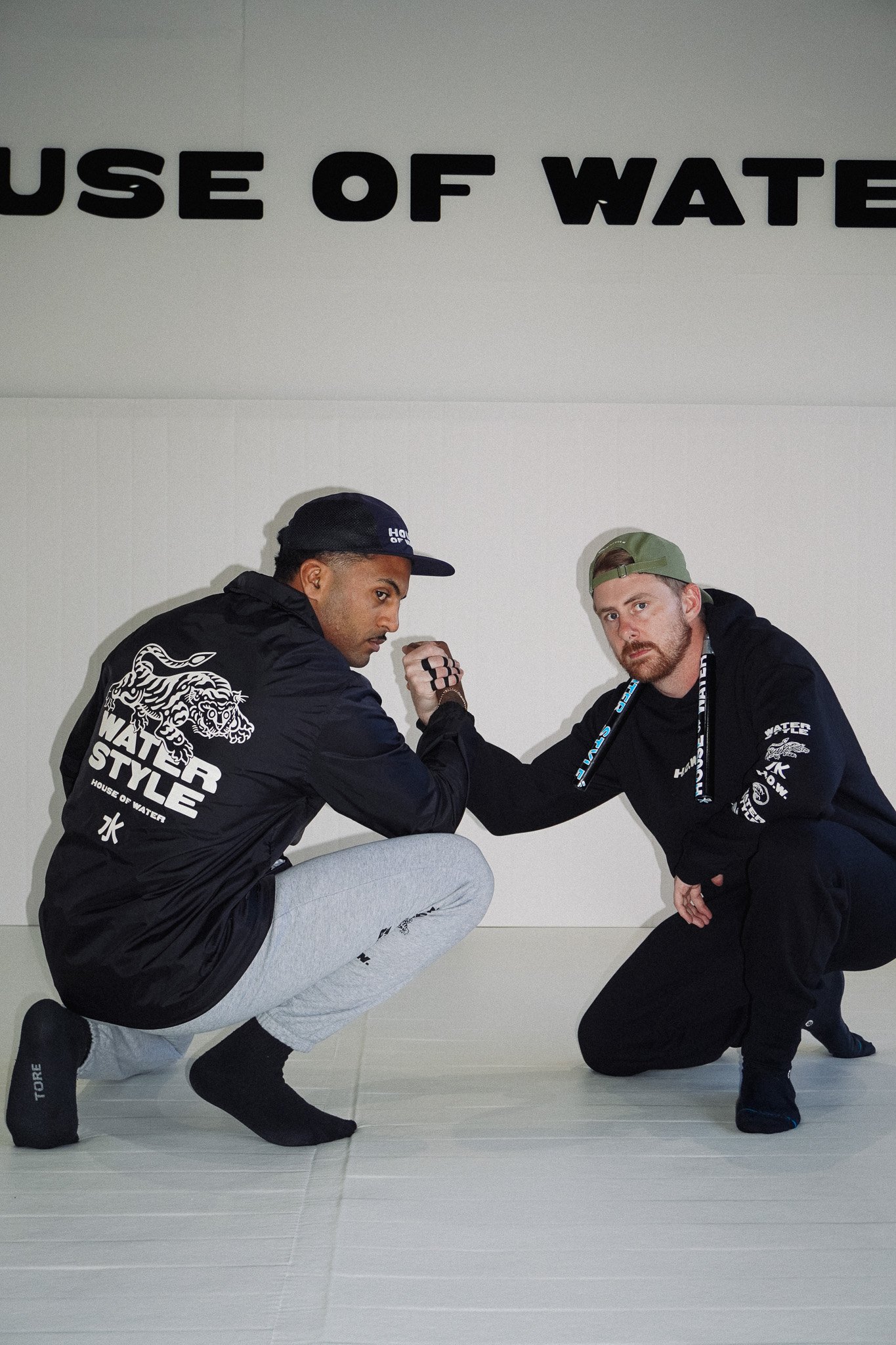 Two men posing in a martial arts studio, arm wrestling, dressed in black hoodies with the House of Water logo, on a white mat with a white wall and partially visible black text 'HOUSE OF WATER' behind them.