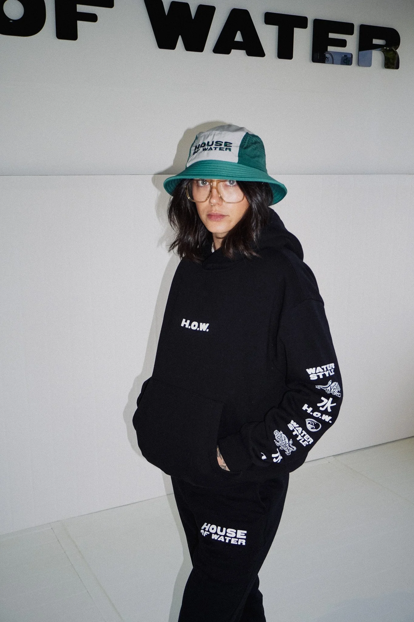 Young woman wearing glasses, a bucket hat, and a black hoodie and pants with white text and graphics, standing against a white wall with the partial words 'HOUSE OF WATER' visible.