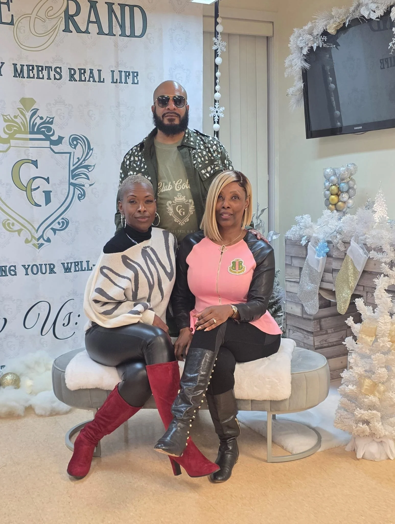 Three people posing in front of a Christmas-themed backdrop with a white Christmas tree decorated with silver and gold ornaments, white stockings, and a banner with the words 'Grand' and 'Real Life'.