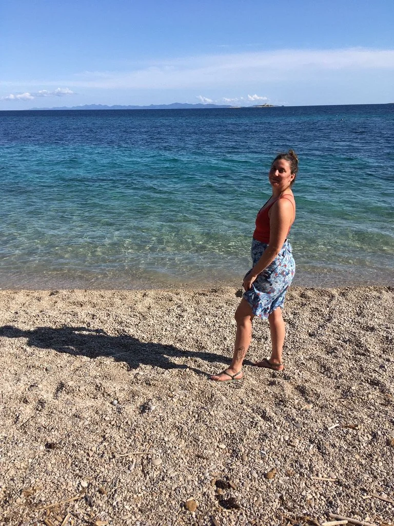 A woman standing on a pebble beach near the ocean with a distant landmass on the horizon, wearing a red top and floral skirt, smiling.
