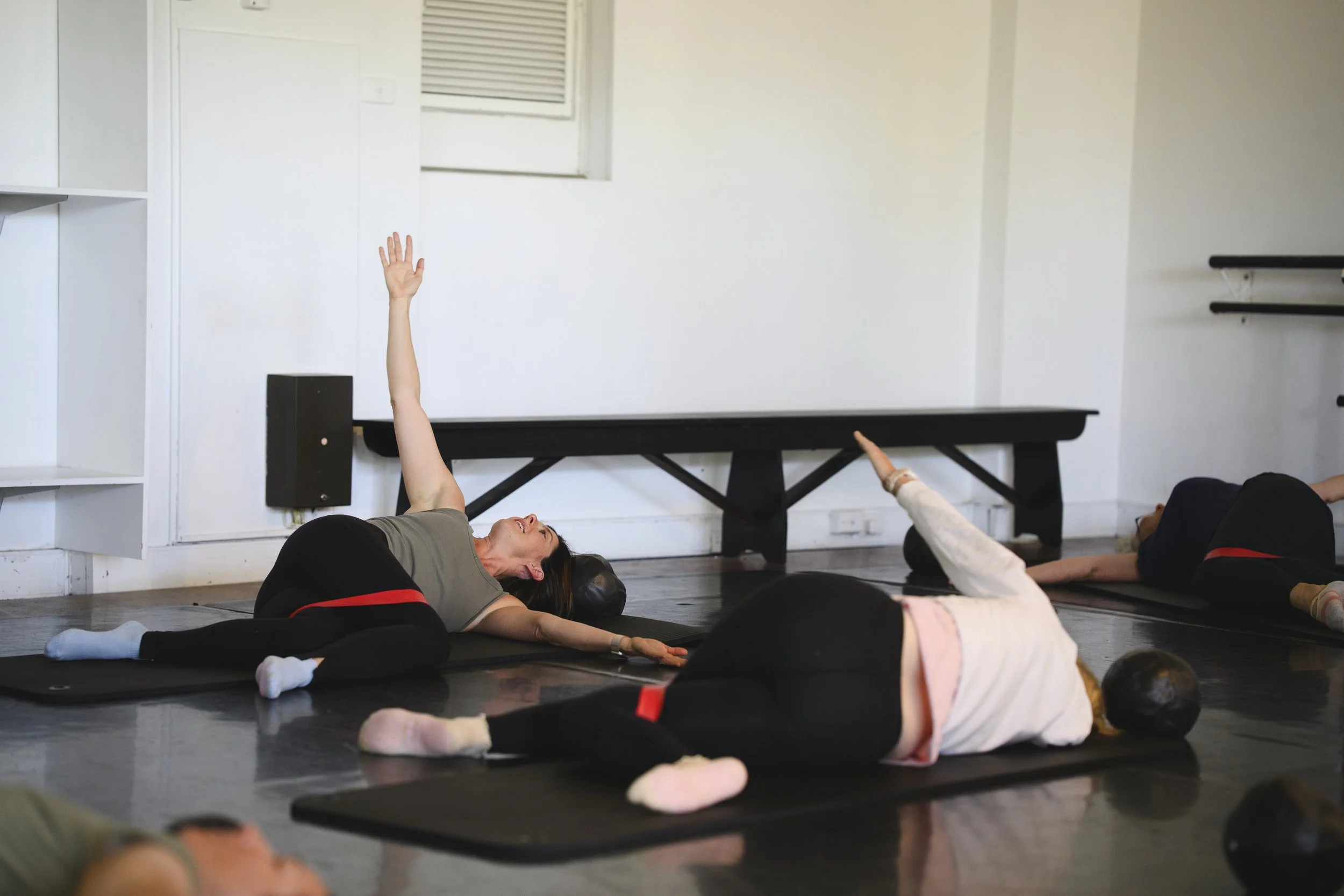 People lying on yoga mats in a Pilates  class, with one woman reaching up with her arm during an exercise in a studio with white walls and a window.