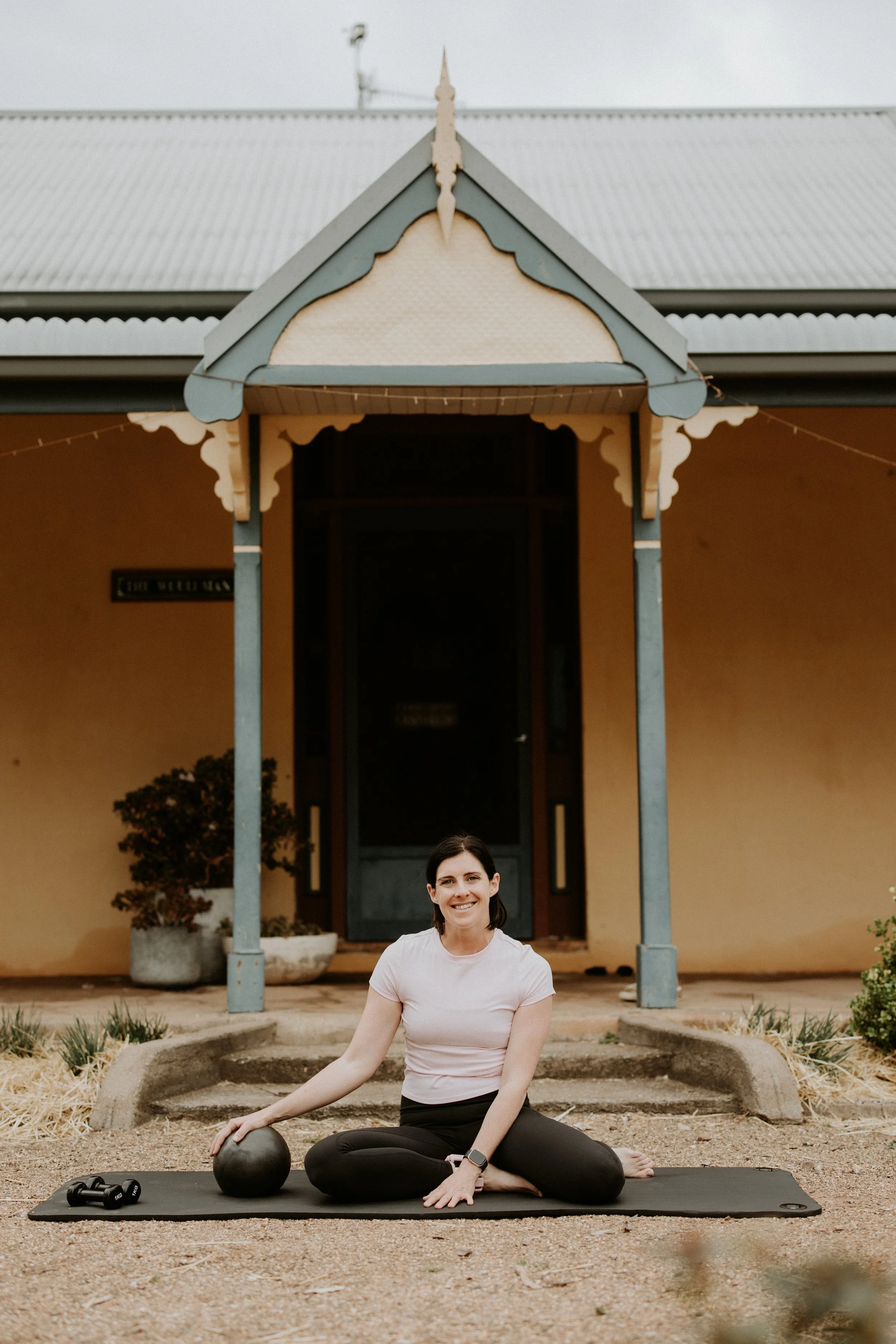 Woman sitting on yoga mat outdoors, smiling, exercise equipment nearby, in front of a building with a porch and decorative trim. Pilates Wellington