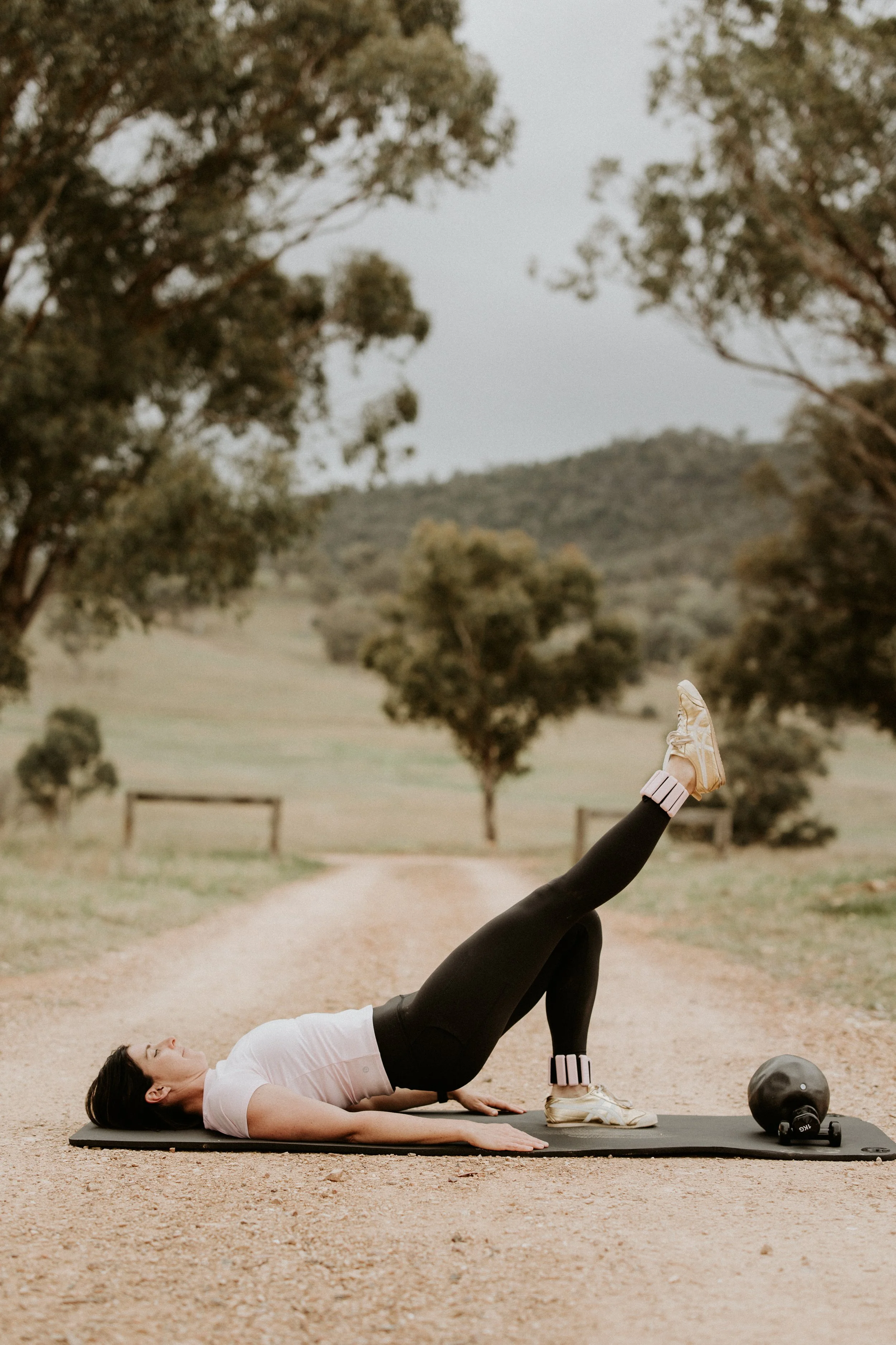 A woman exercising outdoors on a dirt path, lying on her back with one leg raised, wearing athletic clothing and sneakers. There is a fitness ball and a small dumbbell nearby on the ground. Pilates wellington
