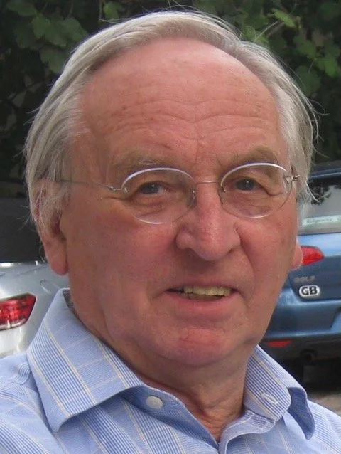 Close-up of an elderly man with glasses and light-colored hair, wearing a light blue collared shirt, outdoors with cars and green foliage in the background.