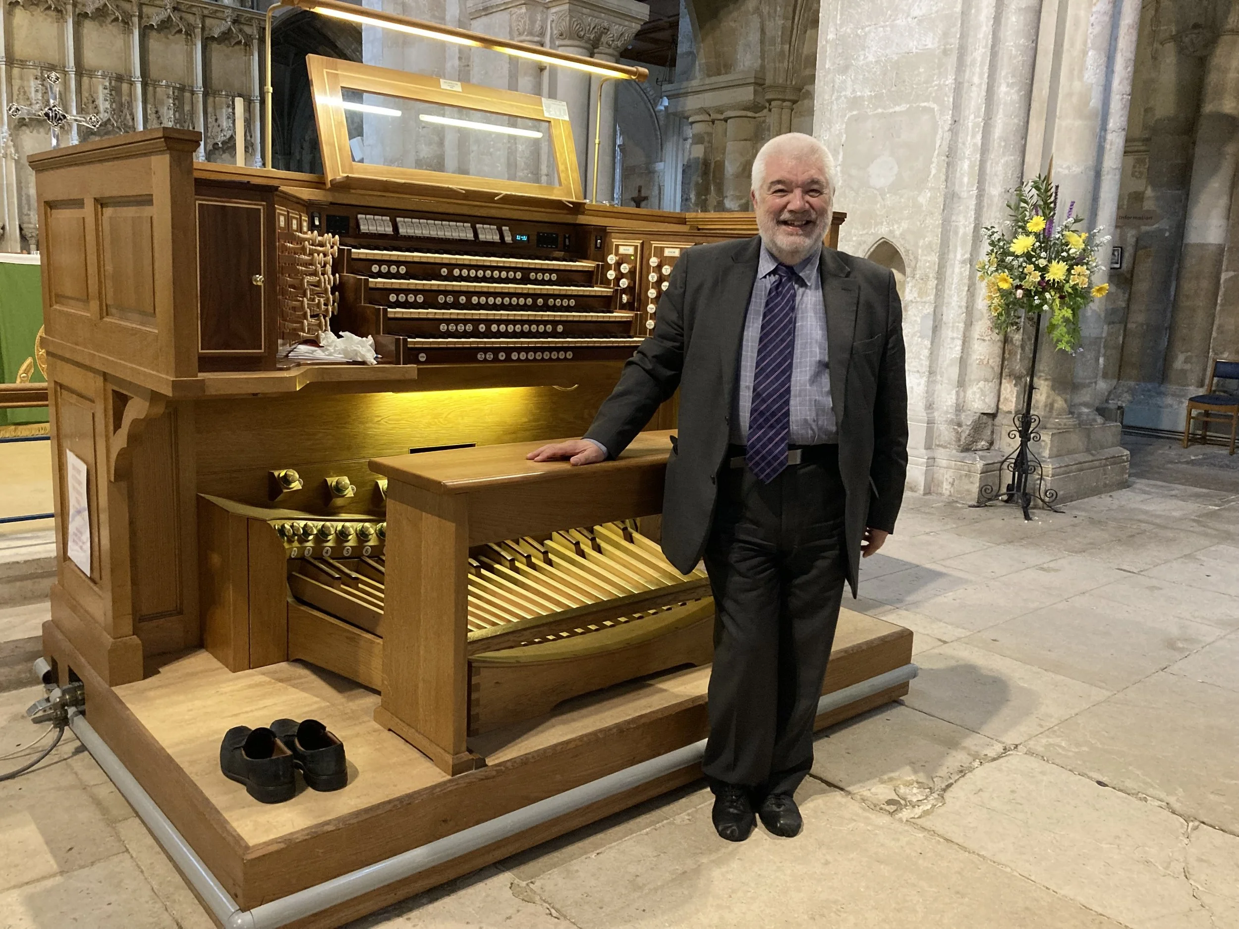 A smiling elderly man in a dark suit, shirt, and tie, standing next to an ornate wooden pipe organ inside a church, with a flower arrangement on a stand nearby.