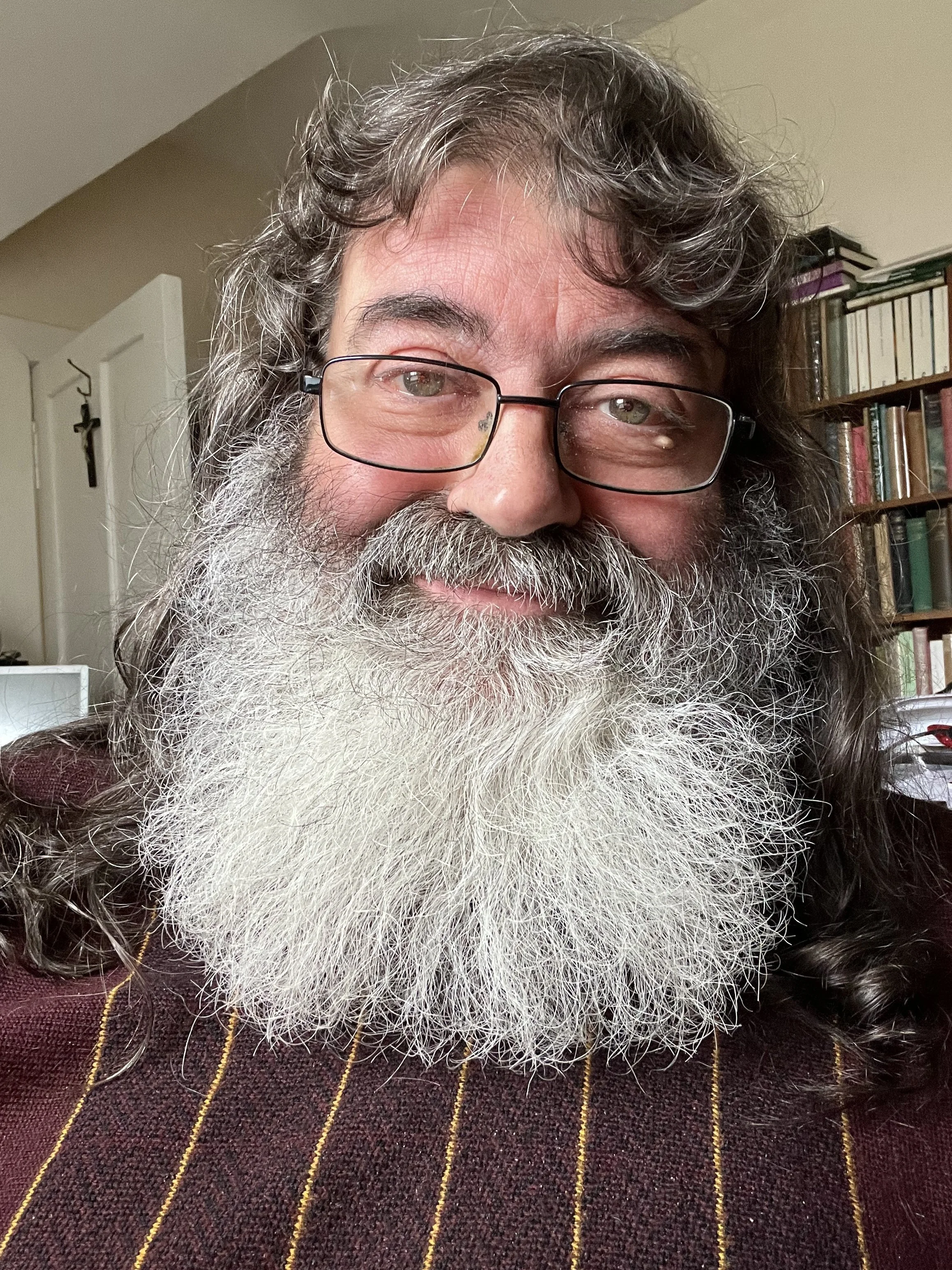 A smiling man with glasses, a full gray beard, and curly gray hair. He is wearing a maroon sweater with yellow stripes. Behind him, there is a bookshelf filled with books and a cross hanging on the wall.