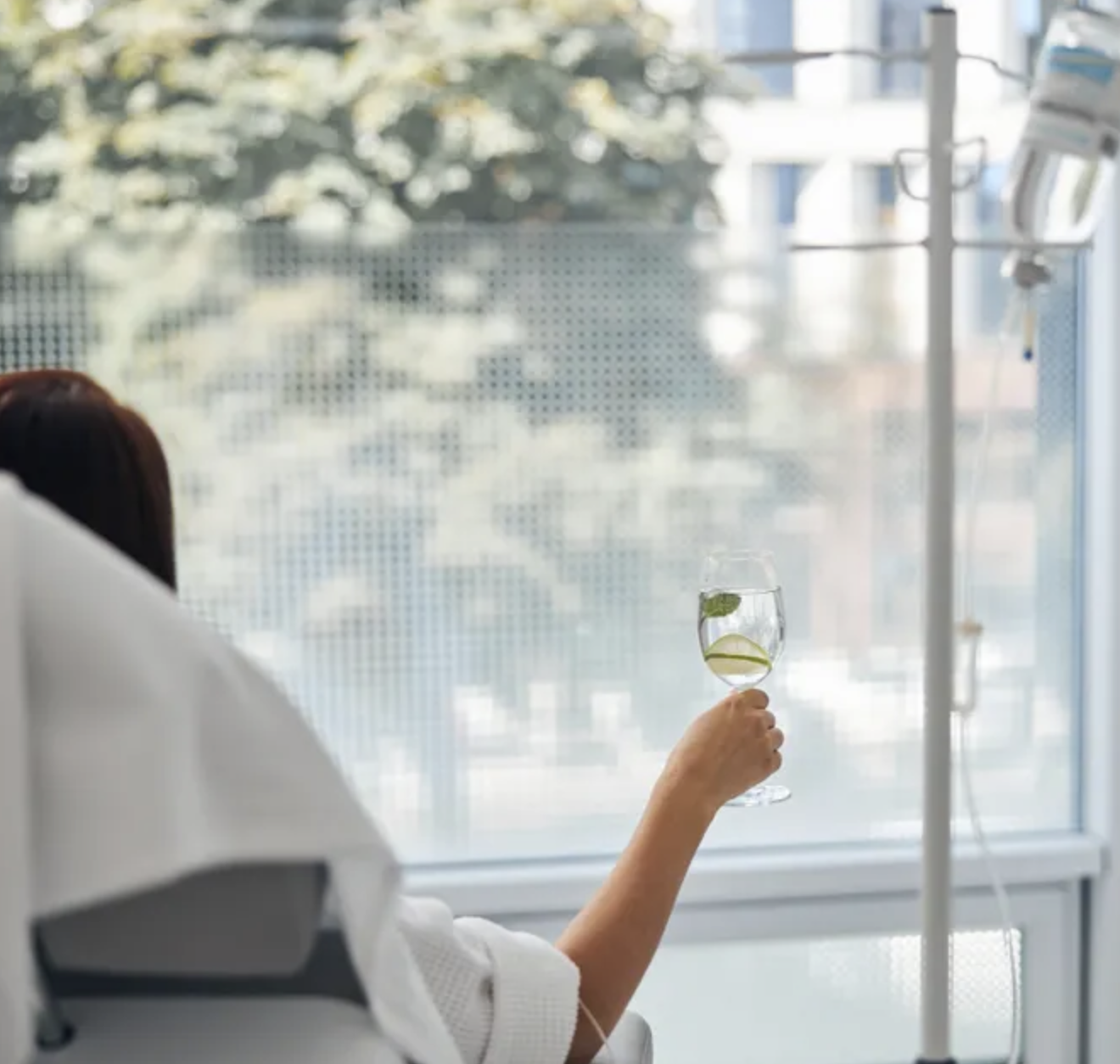 A person sitting in a hospital room holding a glass of water with lemon and mint slices, with IV poles and medication on the right side, and a window with trees outside in the background.