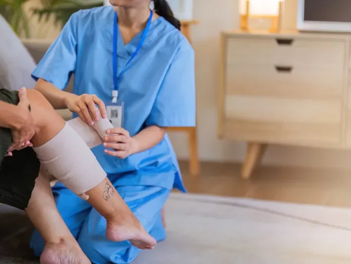 Nurse in blue scrubs checking patient's leg with bandage in a medical room.