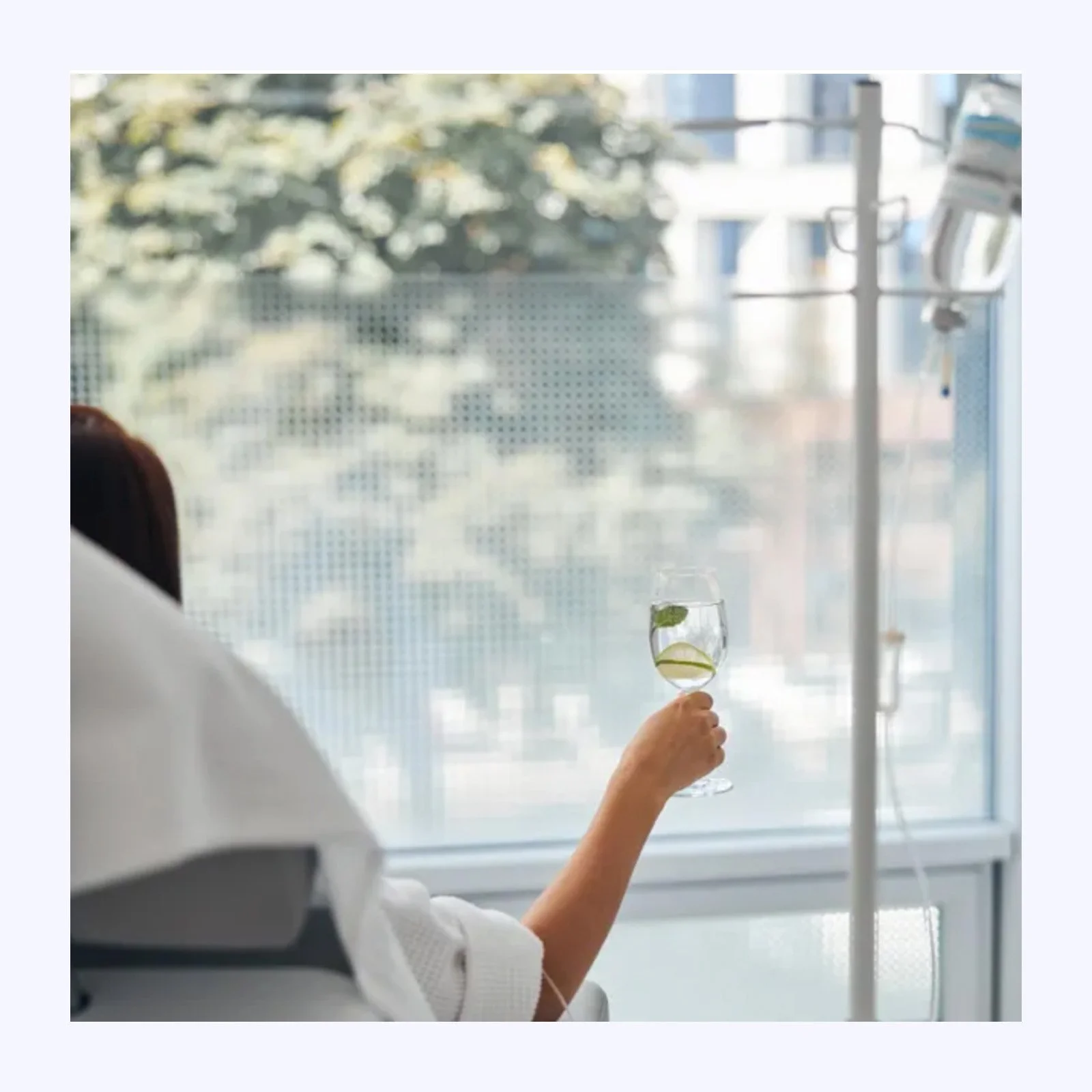 A person sitting in a hospital room holding a glass of water with lemon and mint slices, with IV poles and medication on the right side, and a window with trees outside in the background.