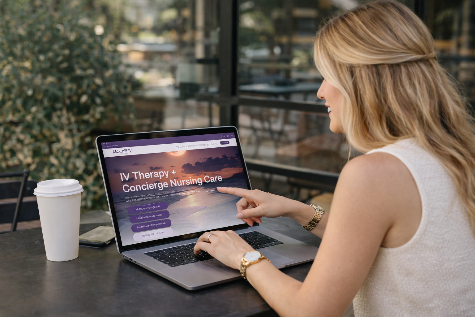 Woman with blonde hair using a laptop outdoors, browsing a website for IV therapy and concierge nursing care, with a coffee cup beside her.