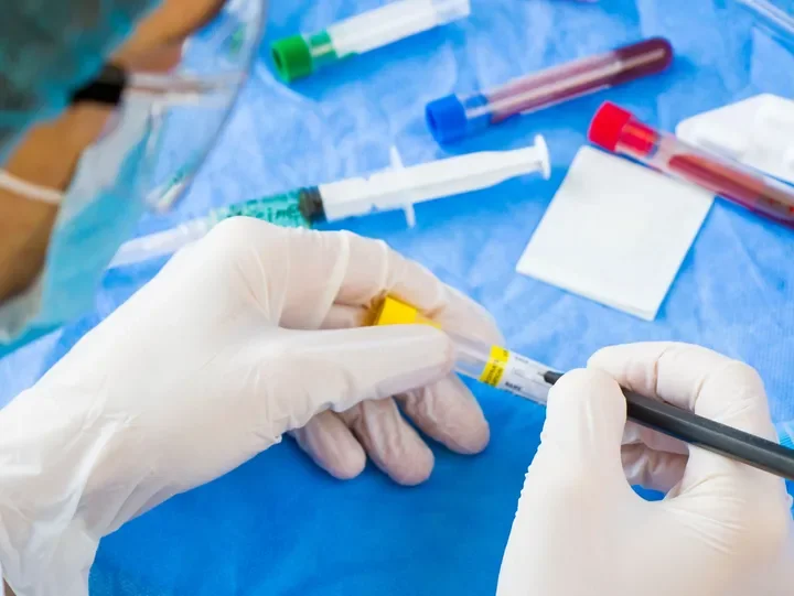 Close-up of healthcare worker's gloved hands preparing a syringe for vaccination, with test tubes and medical supplies on a blue surface in the background.