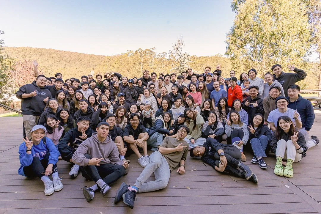 A large group of young adults and teenagers gathered outdoors on a wooden deck, smiling and posing for a group photo with trees and hills in the background on a clear day.