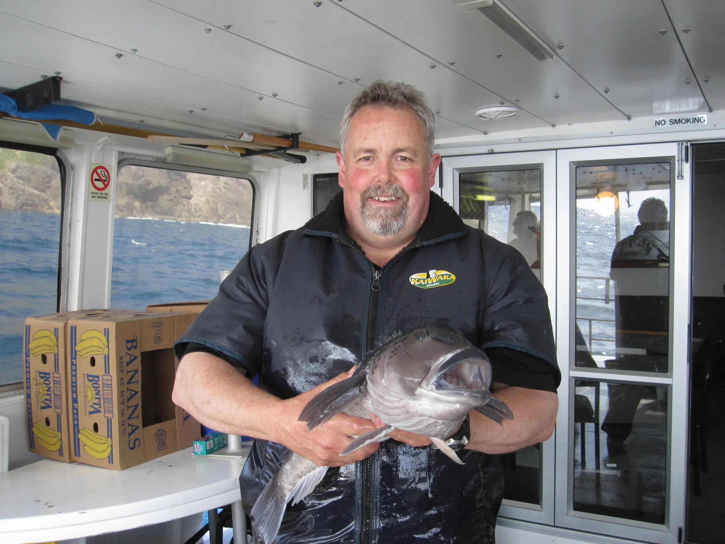 A man holding a large fish inside a boat, with water and rocky cliffs in the background.