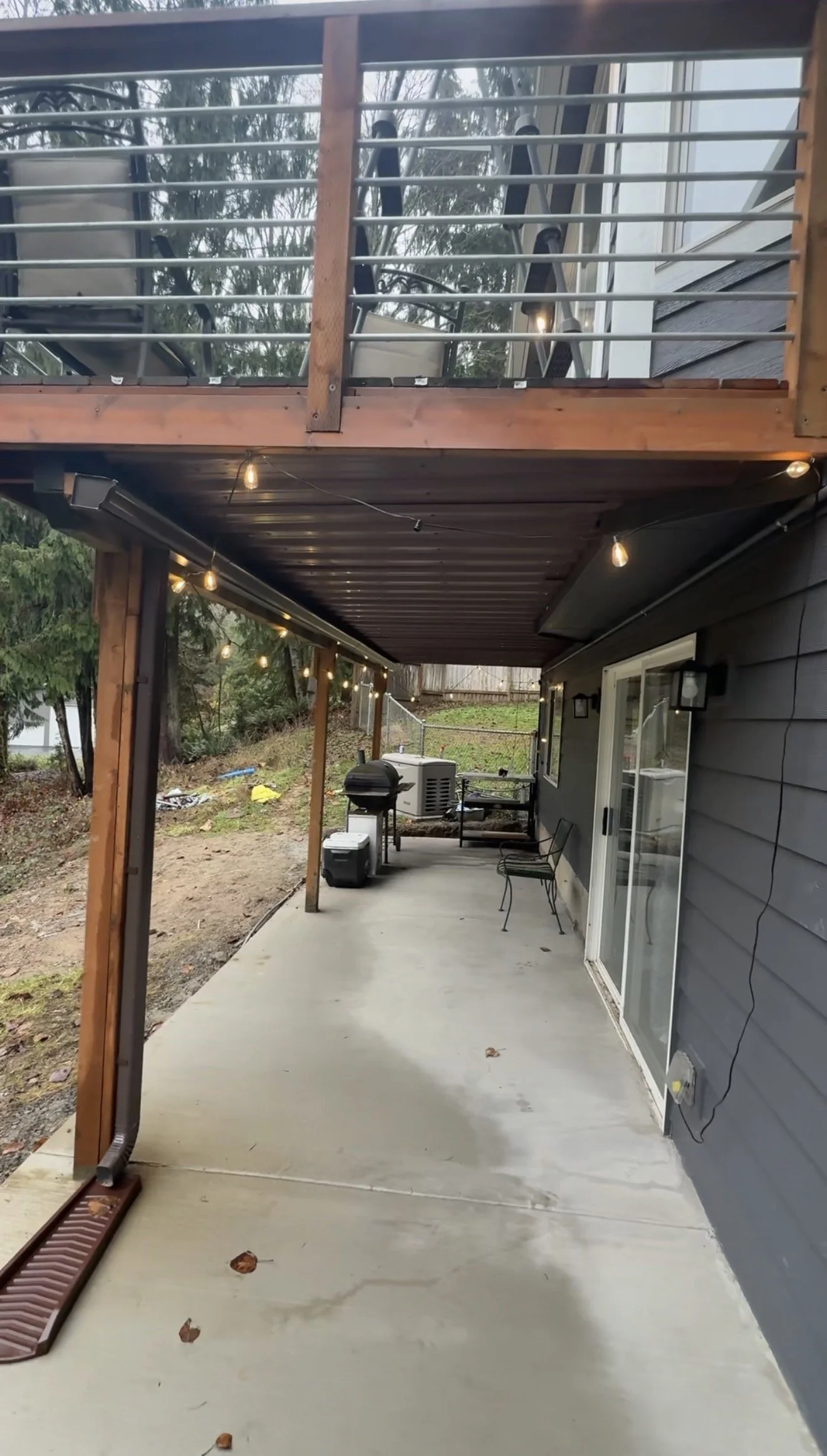 A covered outdoor patio area with a concrete floor, string lights, a grill, and some chairs, attached to a house with gray siding and a sliding glass door. There is a two-story balcony above with outdoor furniture.