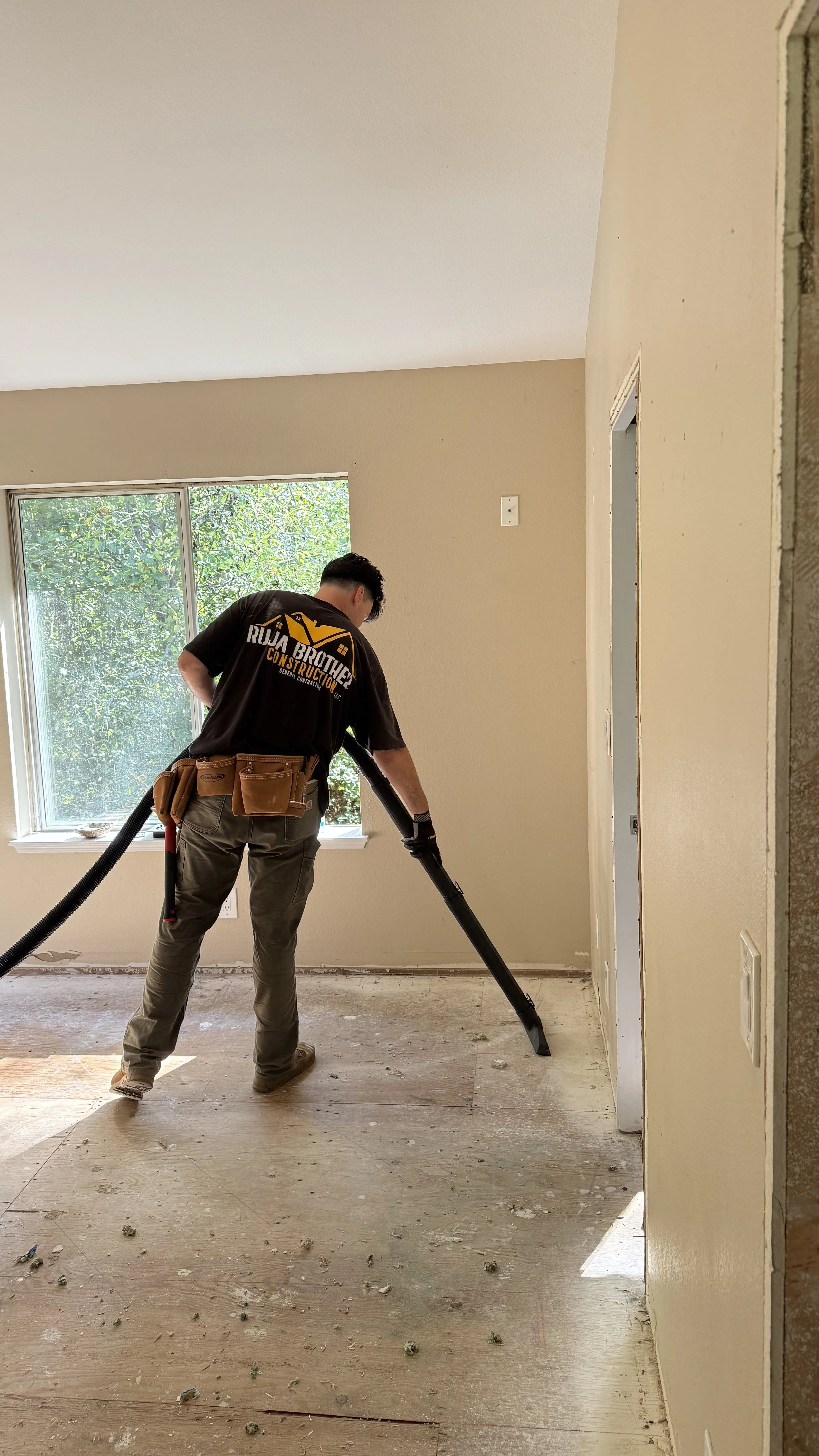 A worker using a vacuum to clean the floor of a room under renovation, with a window showing greenery outside.