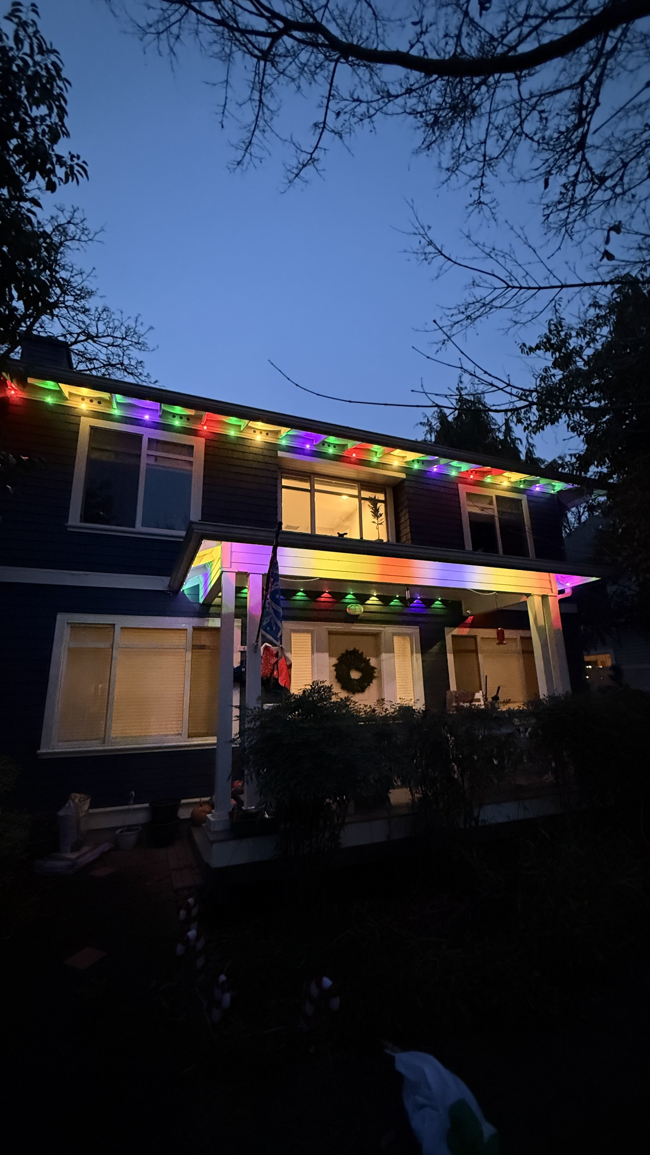 A two-story house decorated with colorful Christmas lights along the roofline and under the porch. The house has multiple windows, a wreath on the front door, and is surrounded by trees and shrubs in the evening.
