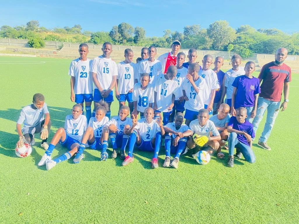 A youth soccer team with coaches on a field, posing for a group photo. The children are wearing white and blue uniforms, some sitting, some standing, with two soccer balls on the ground.