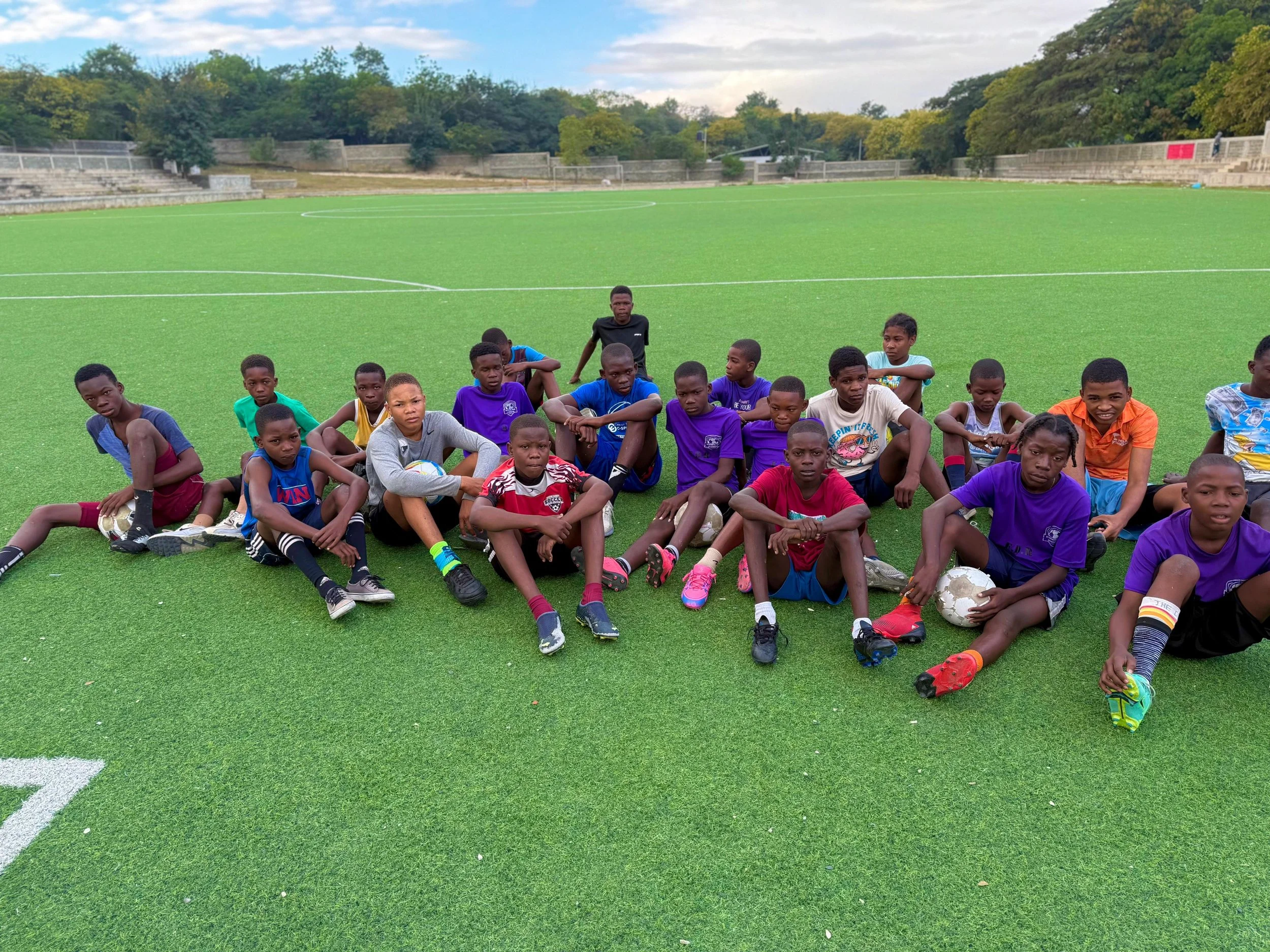 Group of children sitting on artificial turf soccer field, some holding soccer balls, with trees and few people visible in the background.