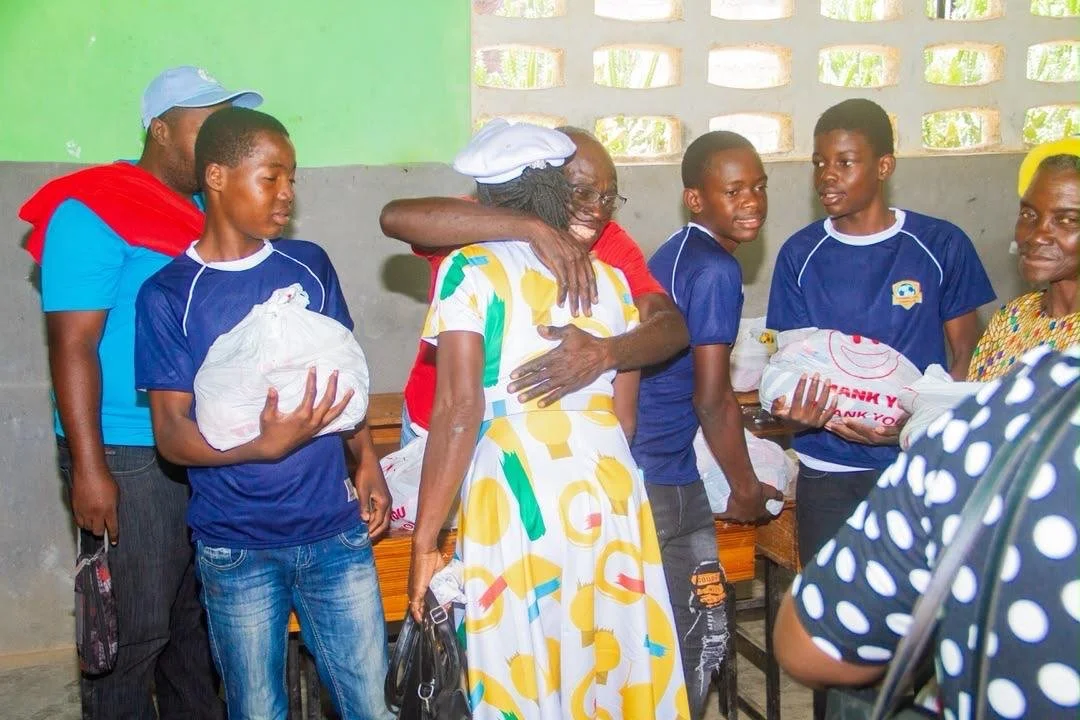A group of people, including children and adults, standing together in an indoor setting, exchanging gifts and hugging each other.