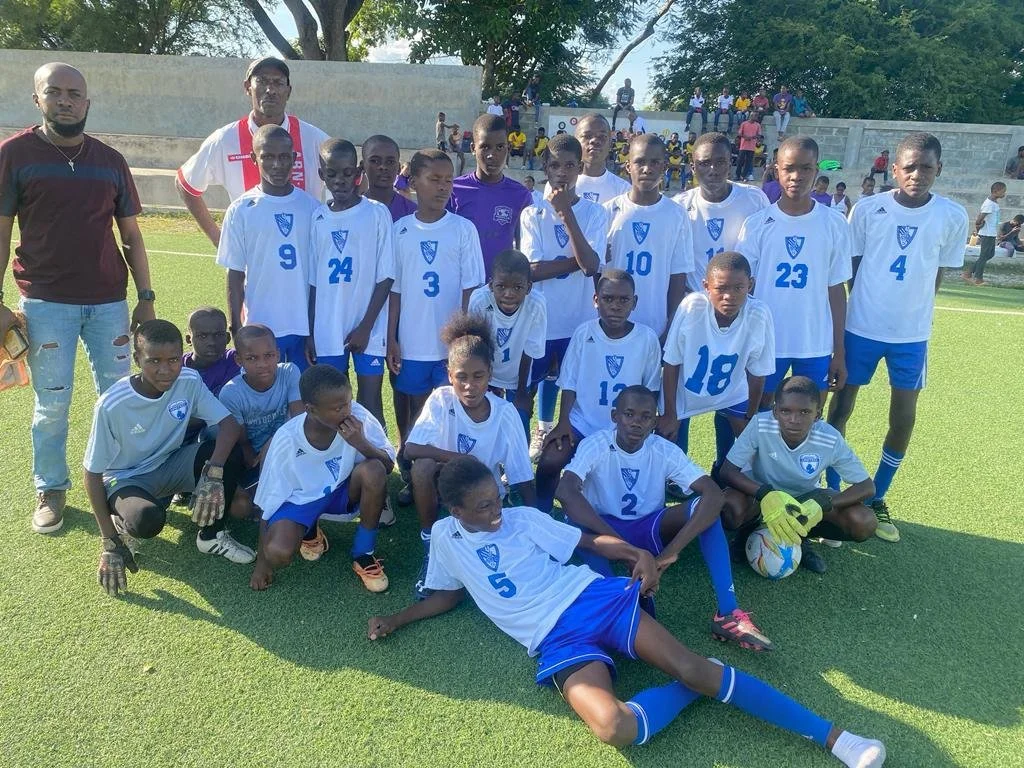 A youth soccer team, along with two adults, standing and sitting on a soccer field for a group photo. The players are wearing white and blue uniforms, and some are holding a soccer ball. Spectators are seated on bleachers in the background.
