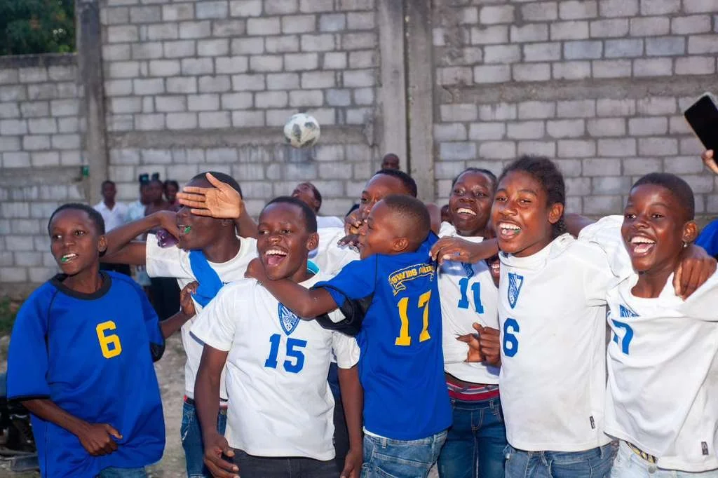 A group of children dressed in soccer jerseys celebrating on a field, with a soccer ball in the air, and smiling.
