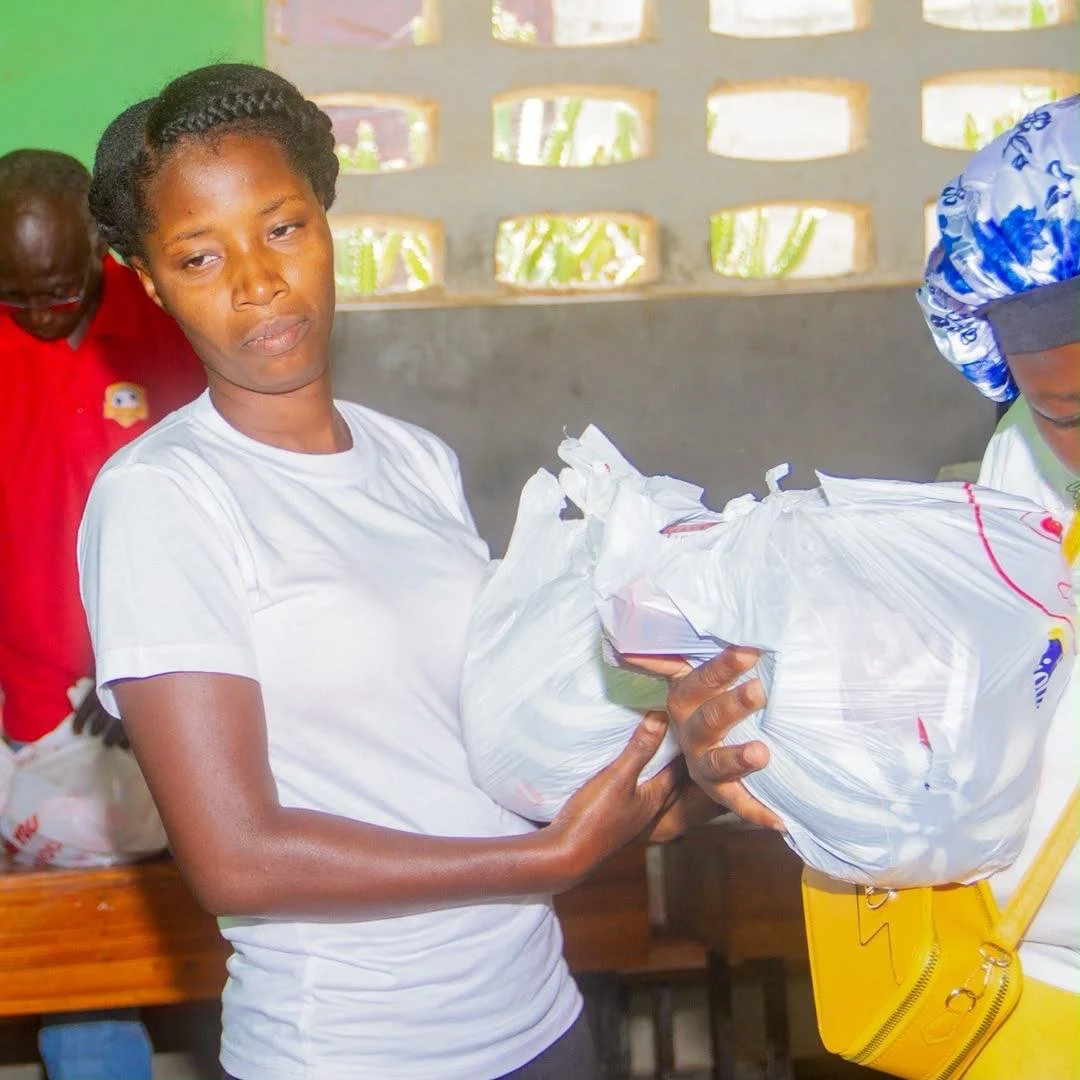 A woman in a white shirt holding a large plastic bag, with a serious facial expression. There is another person in a red shirt in the background and a yellow bag on the right side of the image. The setting appears to be a room with holes in the wall 