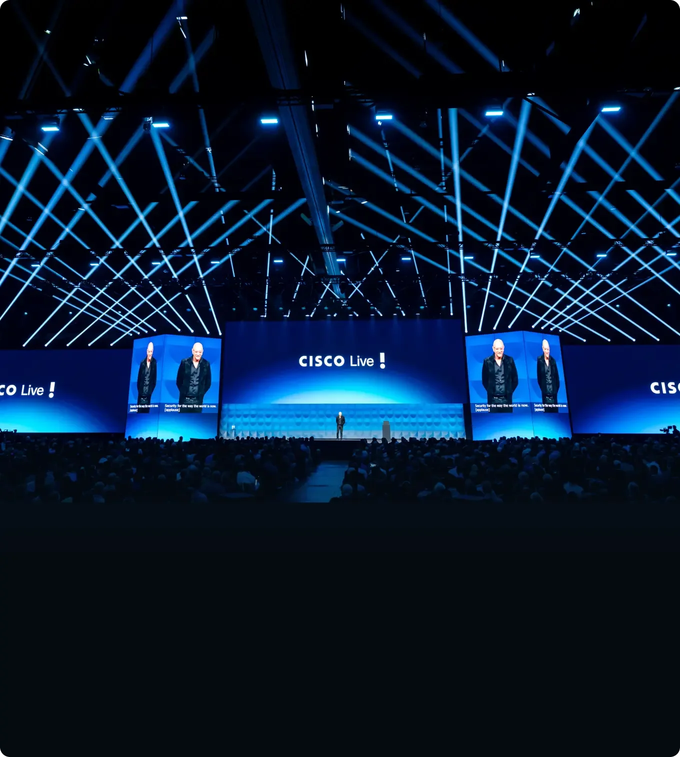 A large conference stage with multiple screens displaying the Cisco logo and speakers, surrounded by a sea of audience members, with blue stage lighting and diagonal light beams overhead.