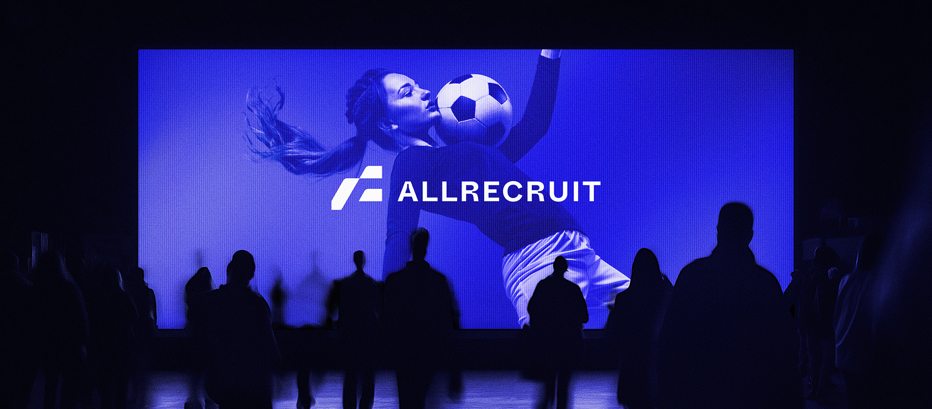 Silhouettes of people watching a large screen displaying the AllRecruit logo and an image of a female soccer player with a ball, in a dark room.
