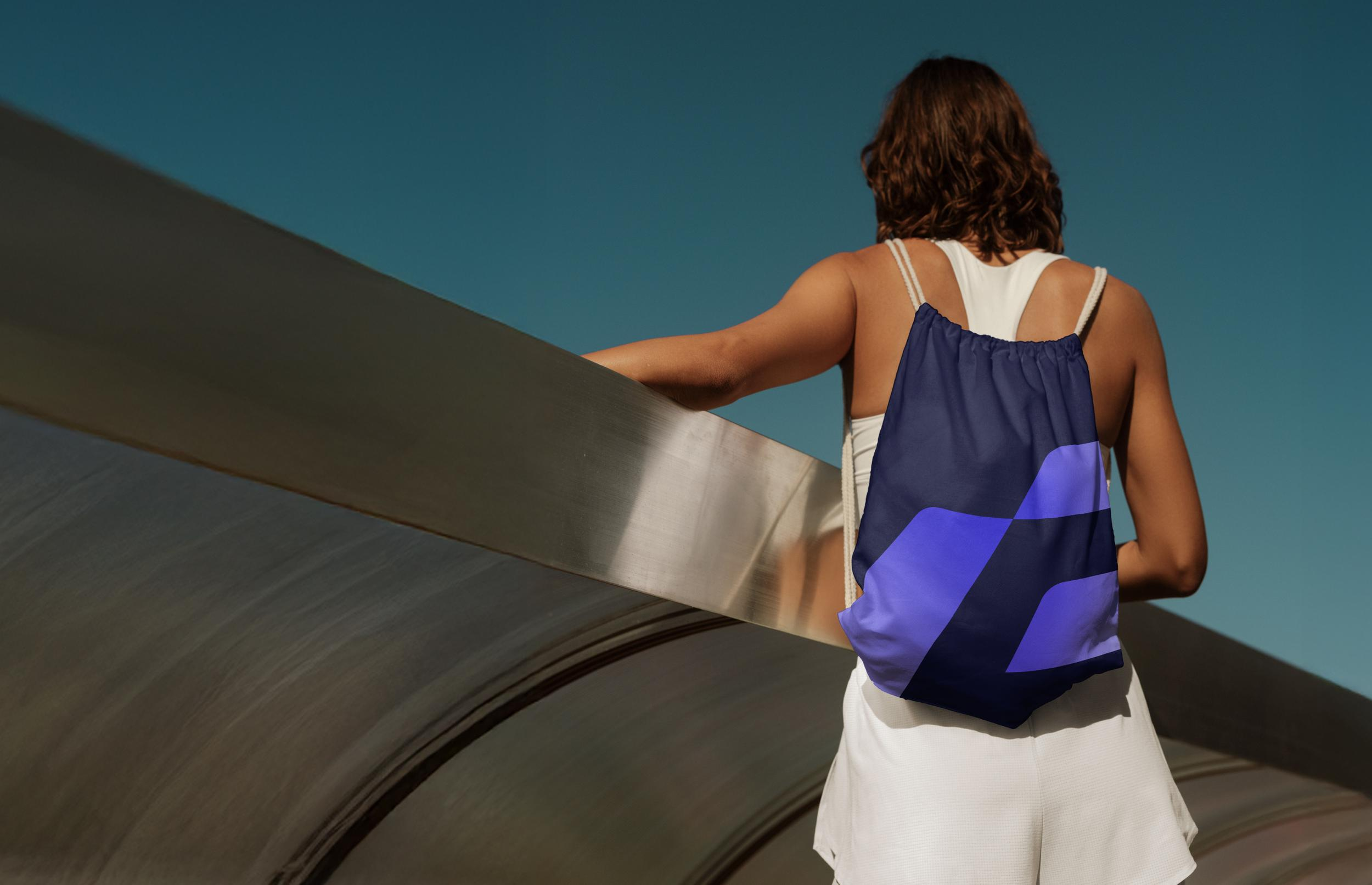 A woman with curly brown hair, wearing a white tank top and white shorts, carries a blue drawstring backpack with geometric design while standing beside a metallic sculpture under a clear blue sky.