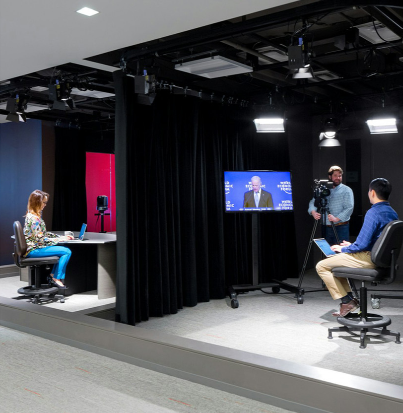 A woman sits at a small round desk with a laptop, participating in a virtual interview or meeting. In the background, a camera operator stands behind a camera on a tripod, filming her. To the right, a man in a blue shirt and beige pants sits with a laptop, possibly hosting or managing the broadcast. A large screen displays a man speaking, likely during a live or recorded presentation. The setting appears to be a professional studio or media room with black curtains and overhead lighting.